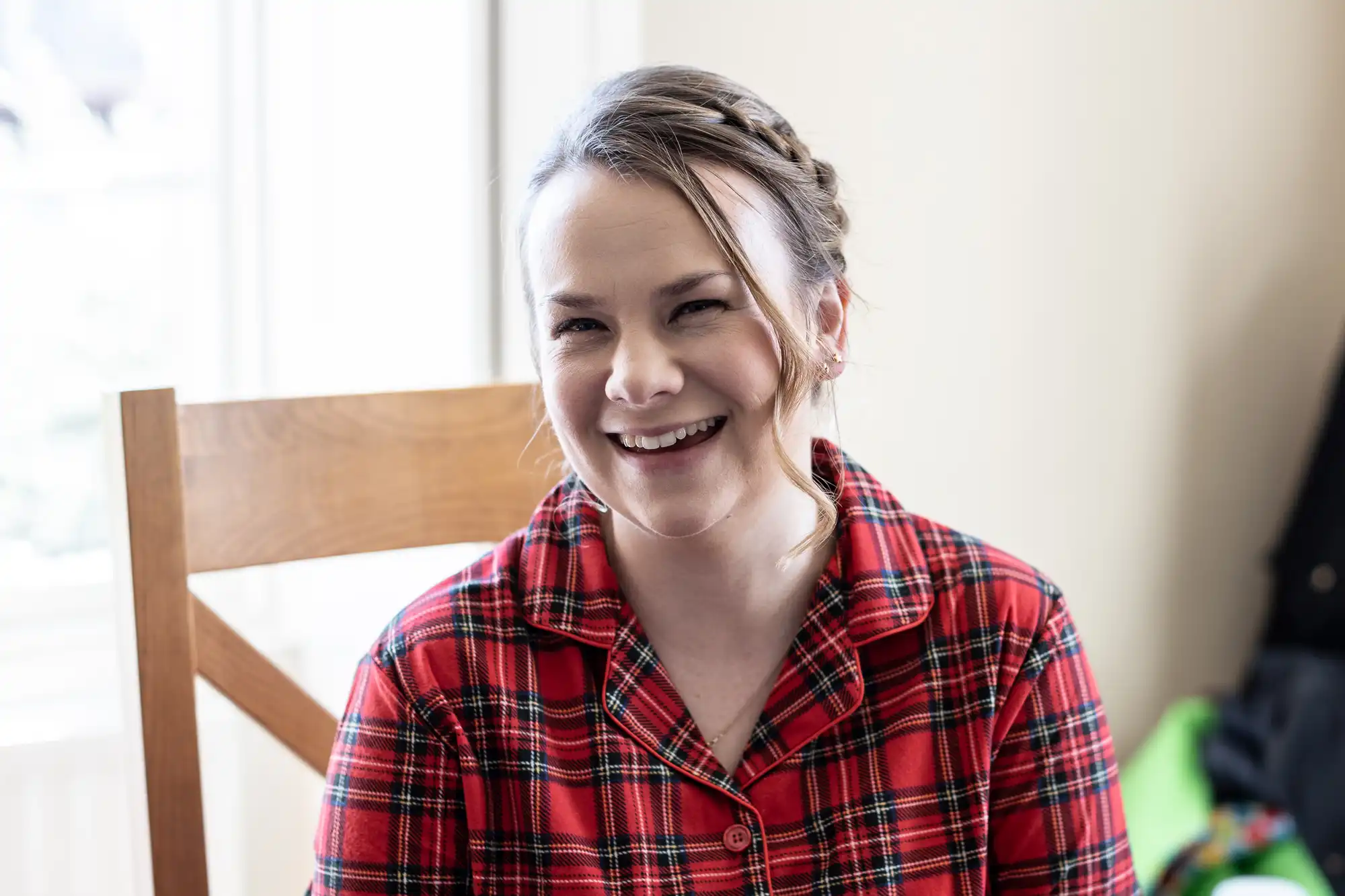 A woman wearing a red plaid shirt is smiling while sitting on a wooden chair in front of a bright window.