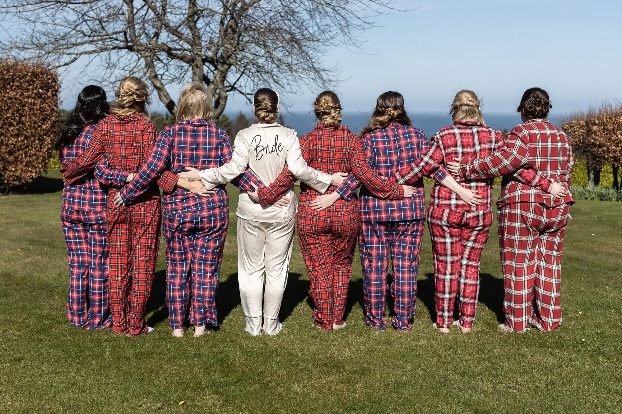 Eight women stand in a row with their backs to the camera, wearing pajamas, with one in white labeled "Bride" and the others in red plaid, in a grassy area with trees and a distant view of water.