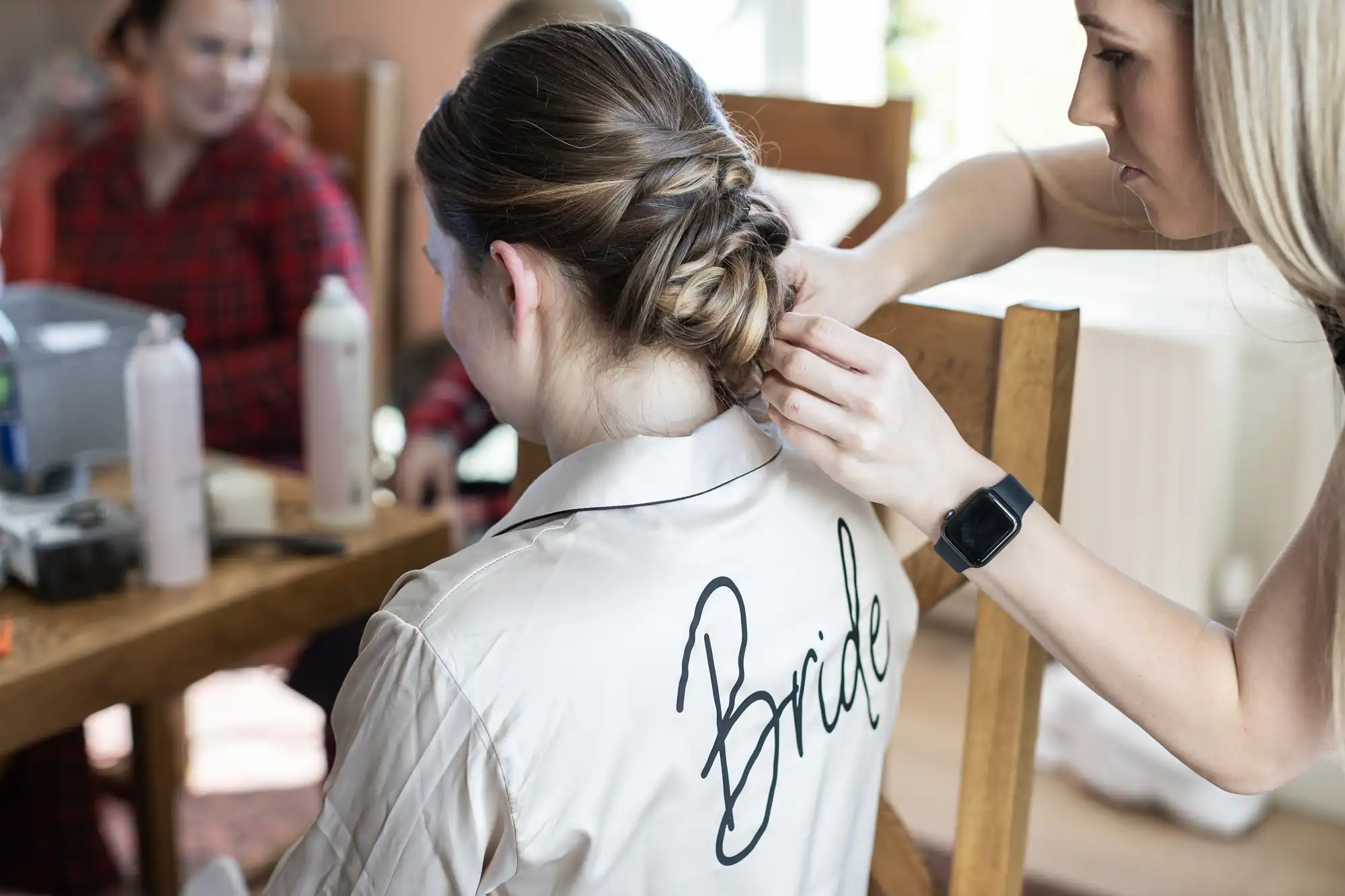 A person styles the hair of a woman wearing a robe with "Bride" written on the back, while others are blurred in the background.