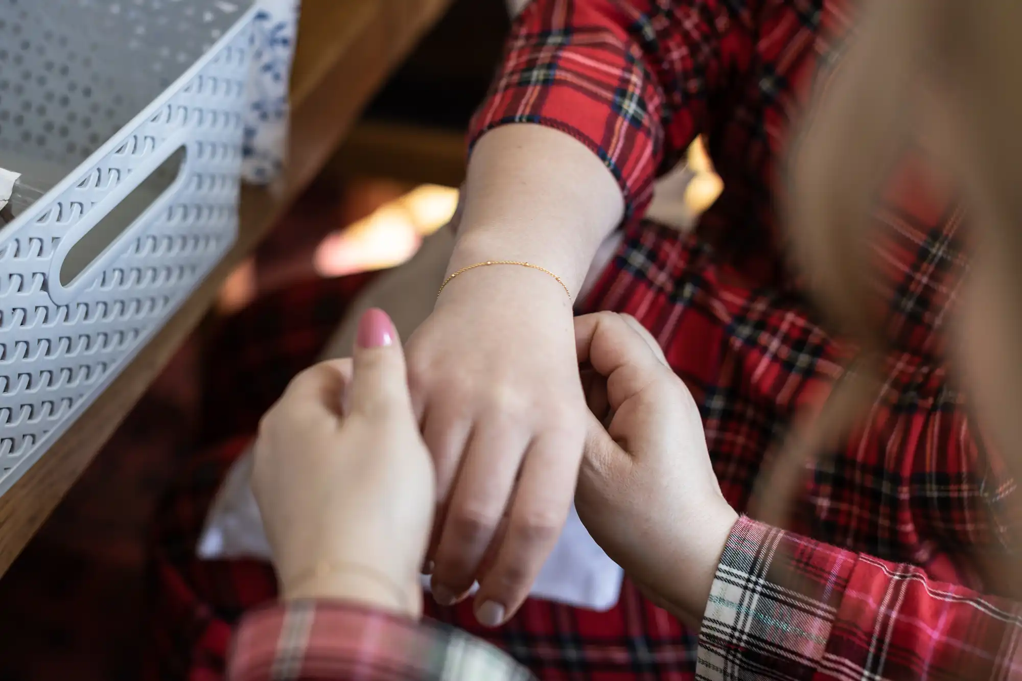 Two people, wearing red plaid clothing, are seen as one attaches a delicate gold bracelet to the wrist of the other. A white woven basket is visible on the table beside them.