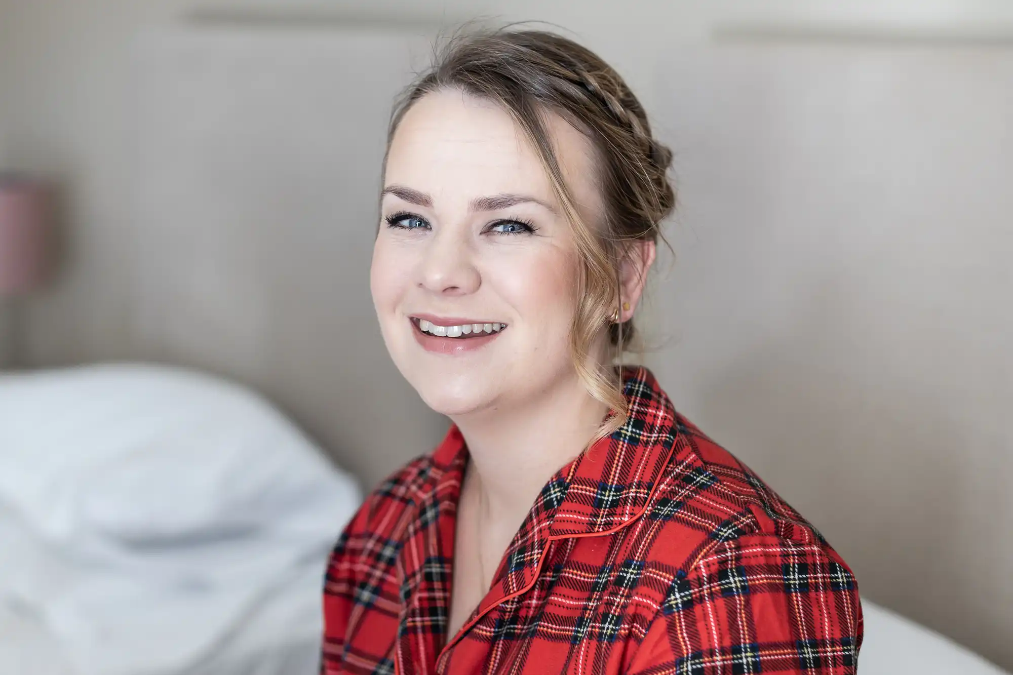 A woman with light-colored hair is smiling while sitting on a bed. She is wearing a red plaid shirt. The background shows a pillow and headboard.