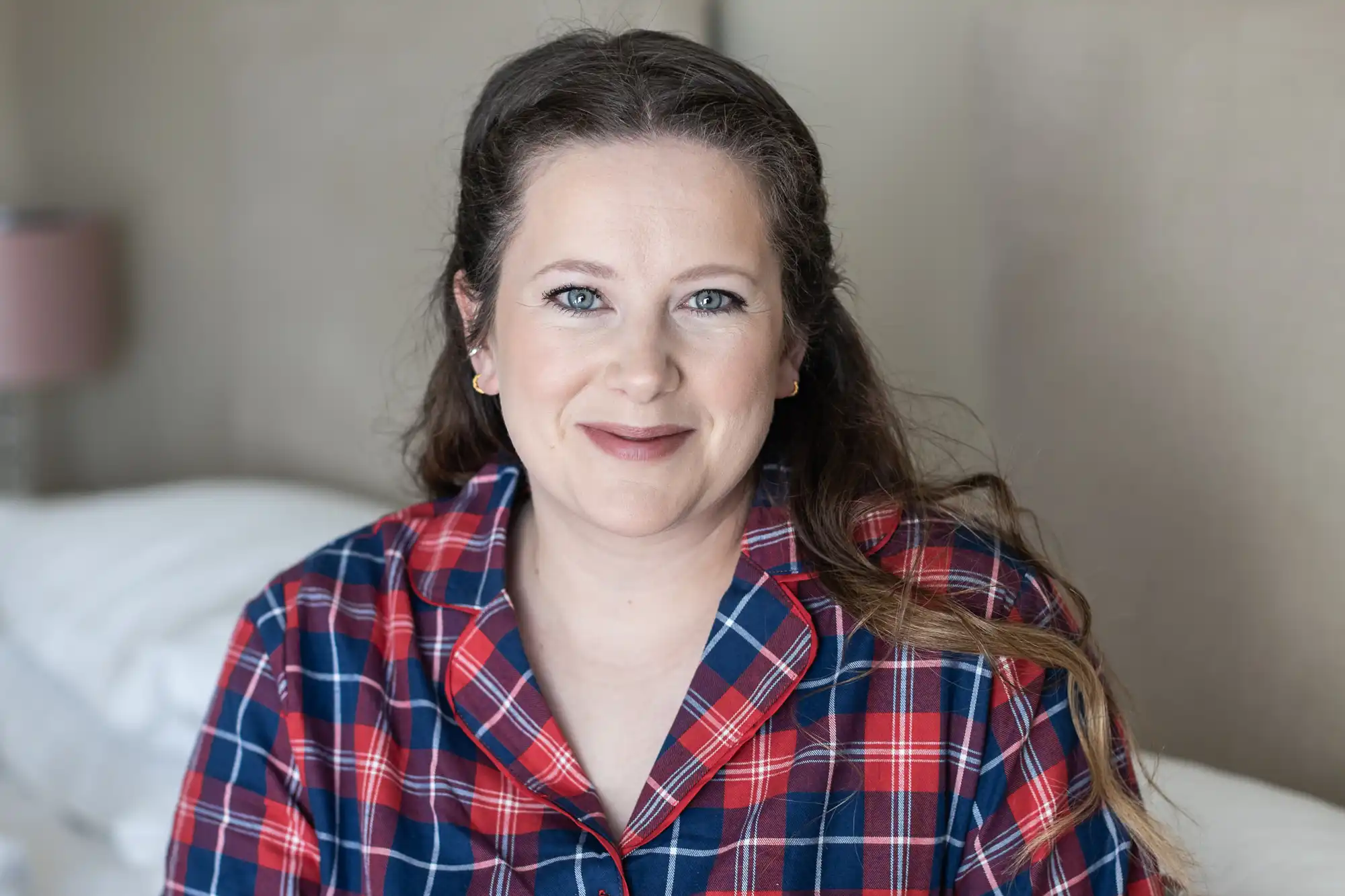 A person with long hair, wearing a red and blue plaid shirt, smiles at the camera while sitting on a bed.