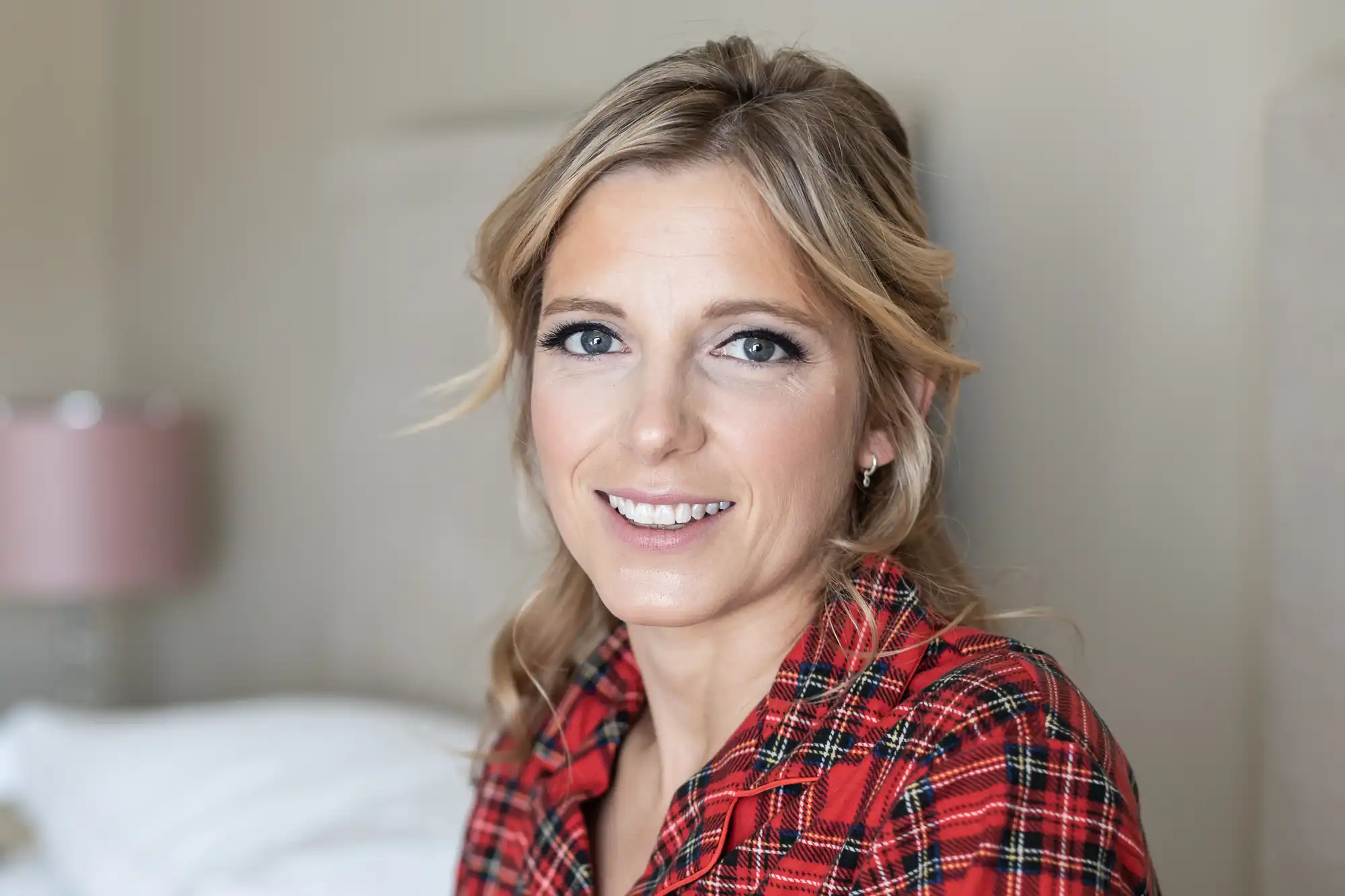 A woman with blonde hair smiles at the camera, wearing a red and black plaid shirt. The background appears to be a bedroom with a bed and a table lamp.