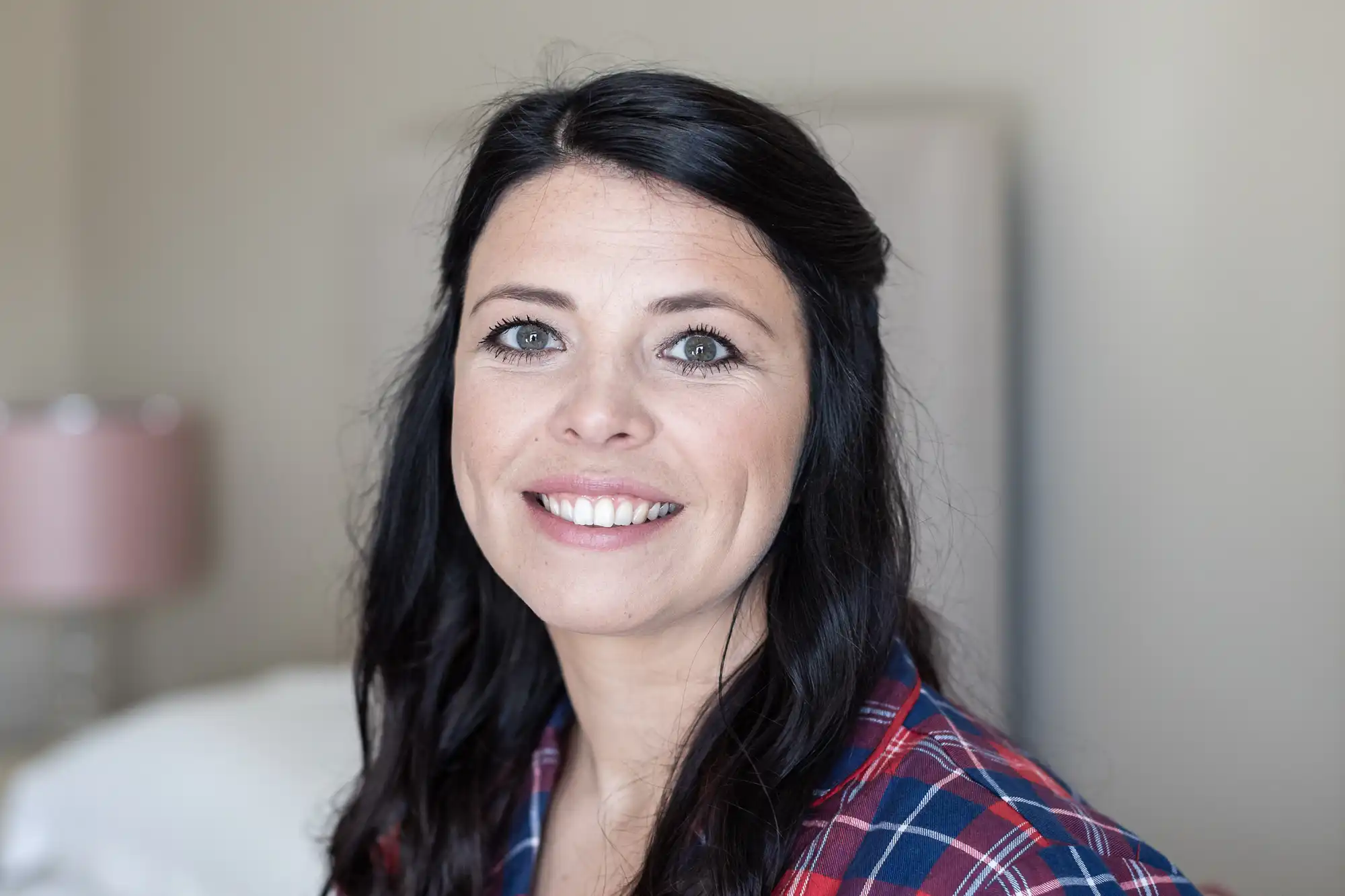 A woman with long dark hair, wearing a plaid shirt, smiles at the camera in an indoor setting.