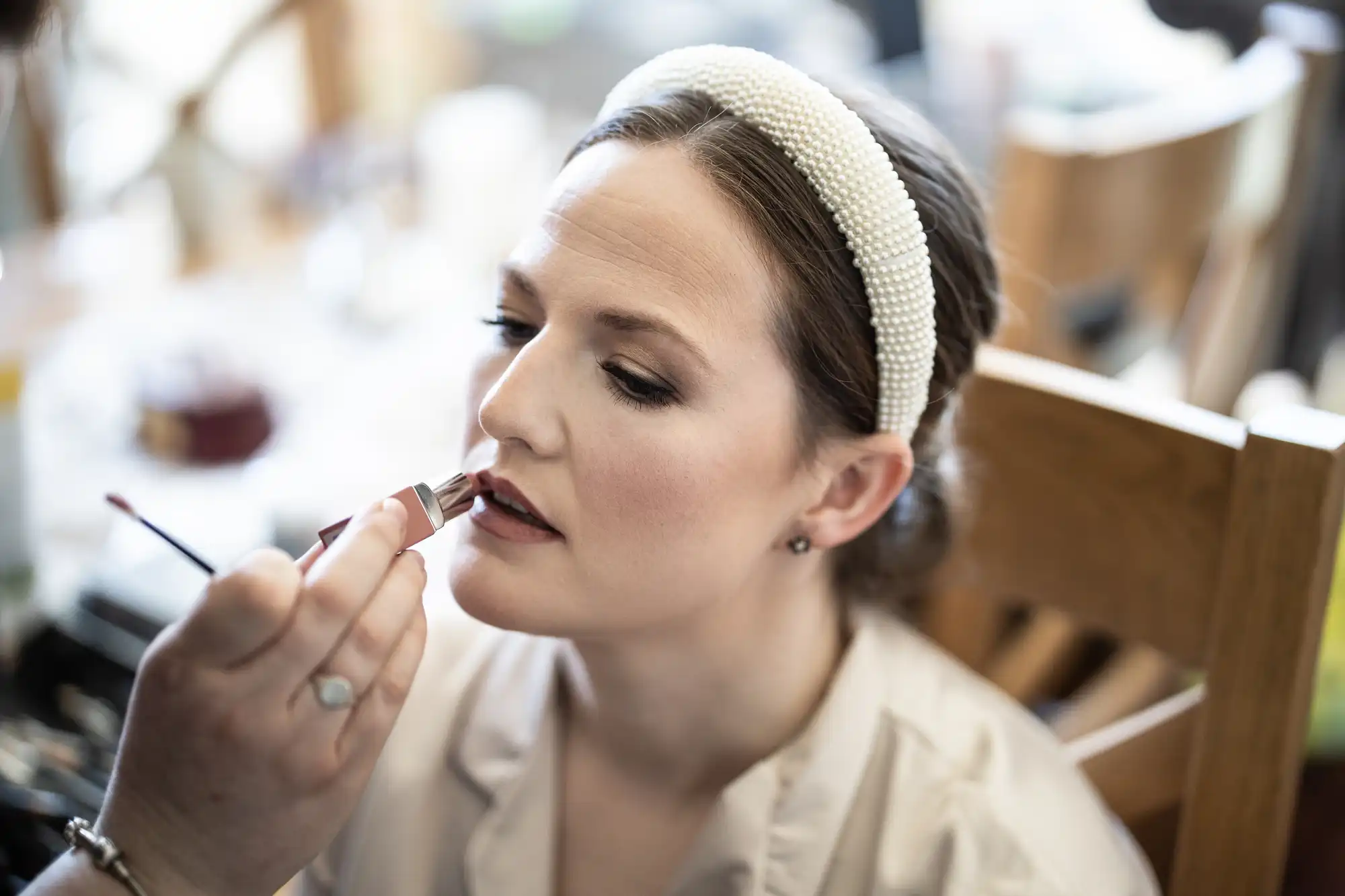 A person wearing a pearl headband is having their lips applied with lipstick by another person.