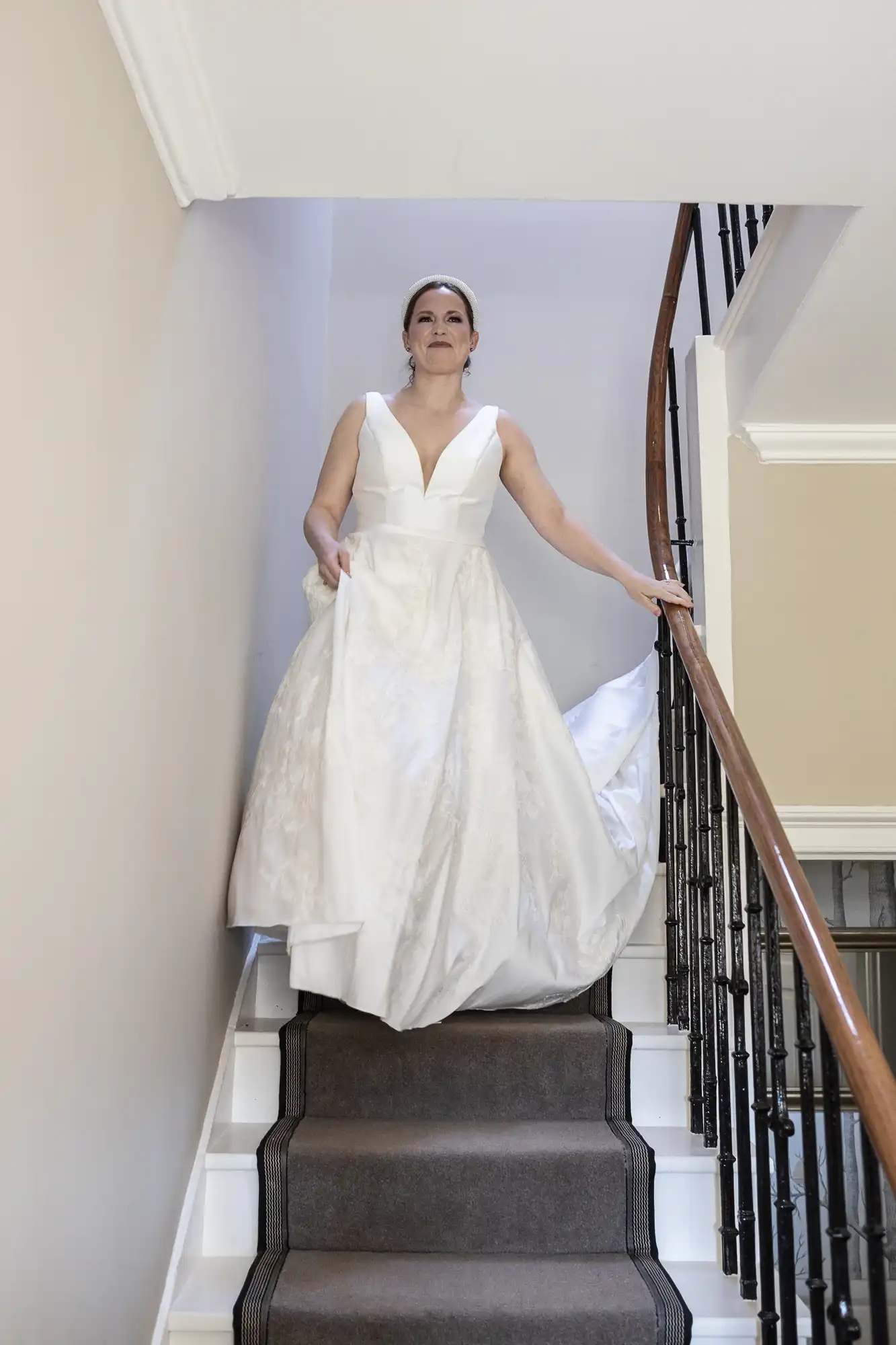 A woman in an elegant white dress descends a curved staircase indoors, holding onto the railing with one hand and lifting her dress with the other.