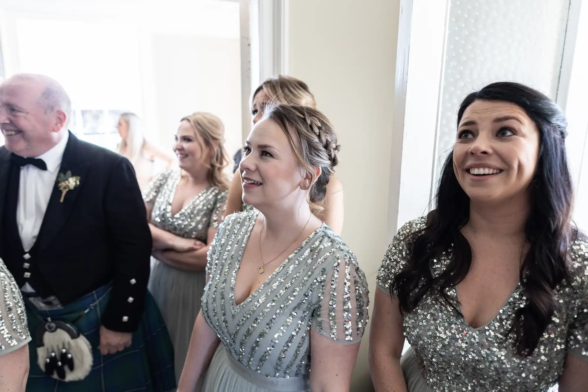 A group of people, including women in silver sequin dresses and a man in a kilt, stand together in a bright room, smiling and looking in one direction.