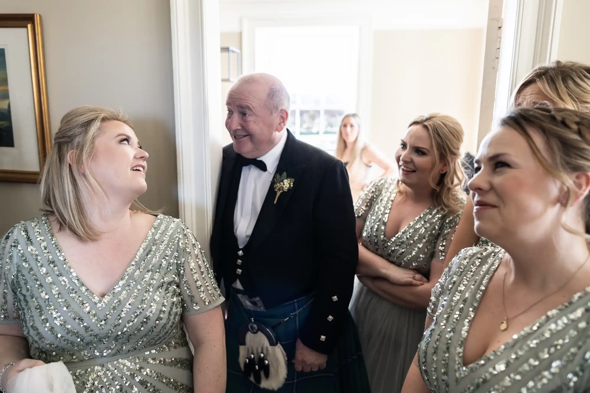 A man in a formal Scottish kilt and jacket smiles while talking to three women in sparkly dresses in a brightly lit room, with a woman in the background.