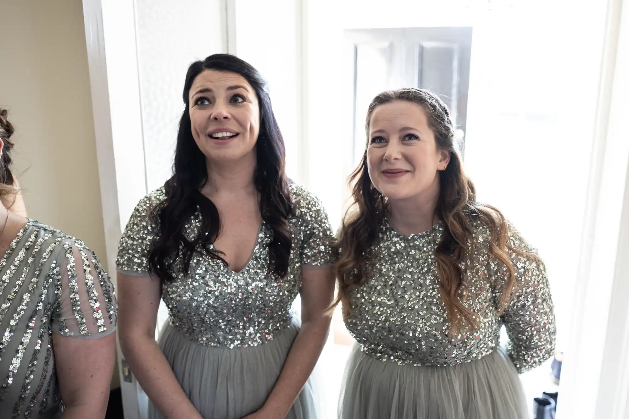 Two women in silver sequin dresses smile while standing indoors, next to a person partially visible on the left.