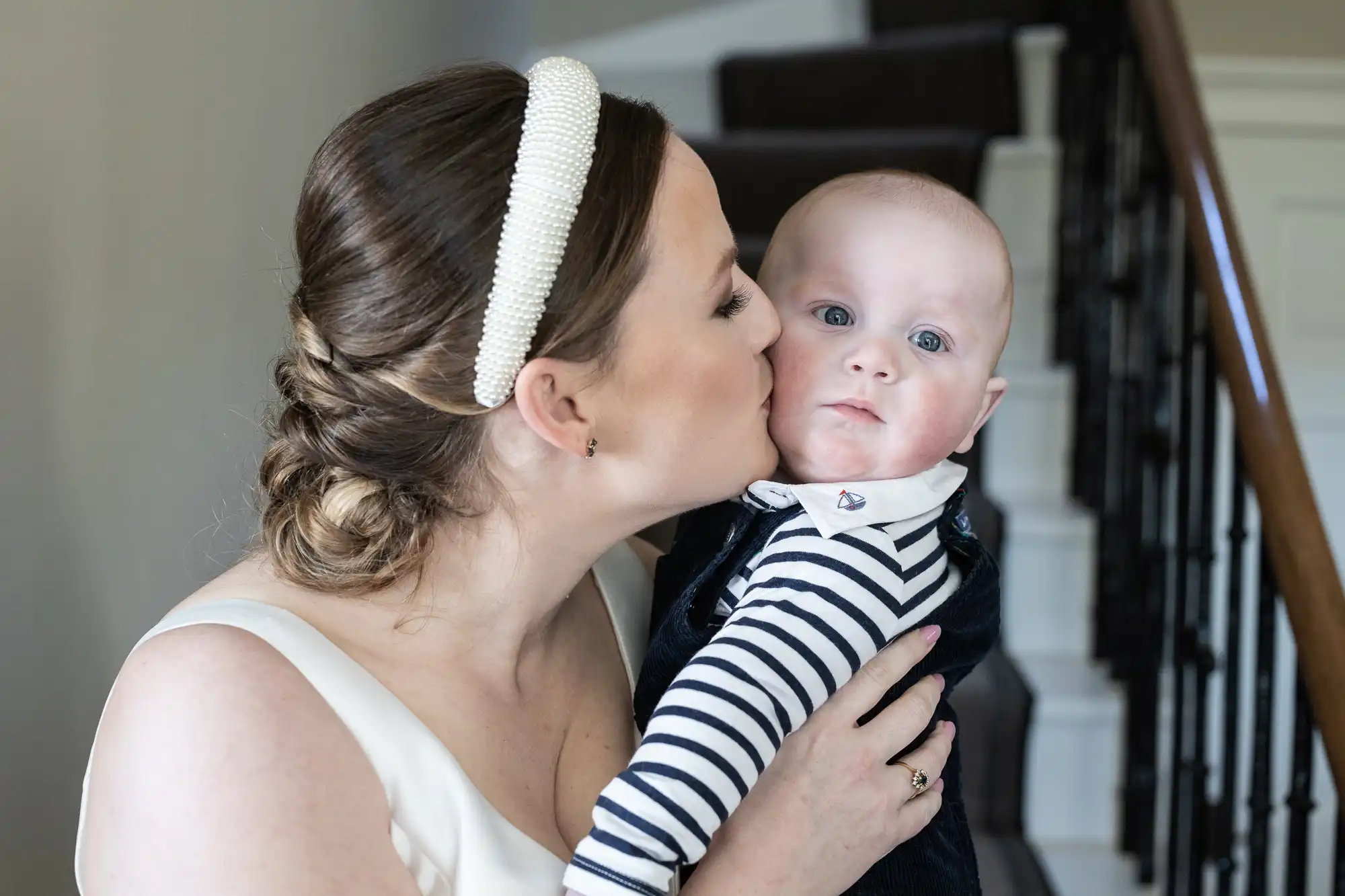 A woman with a beaded headband kisses a baby on the cheek while holding the baby in front of a staircase.