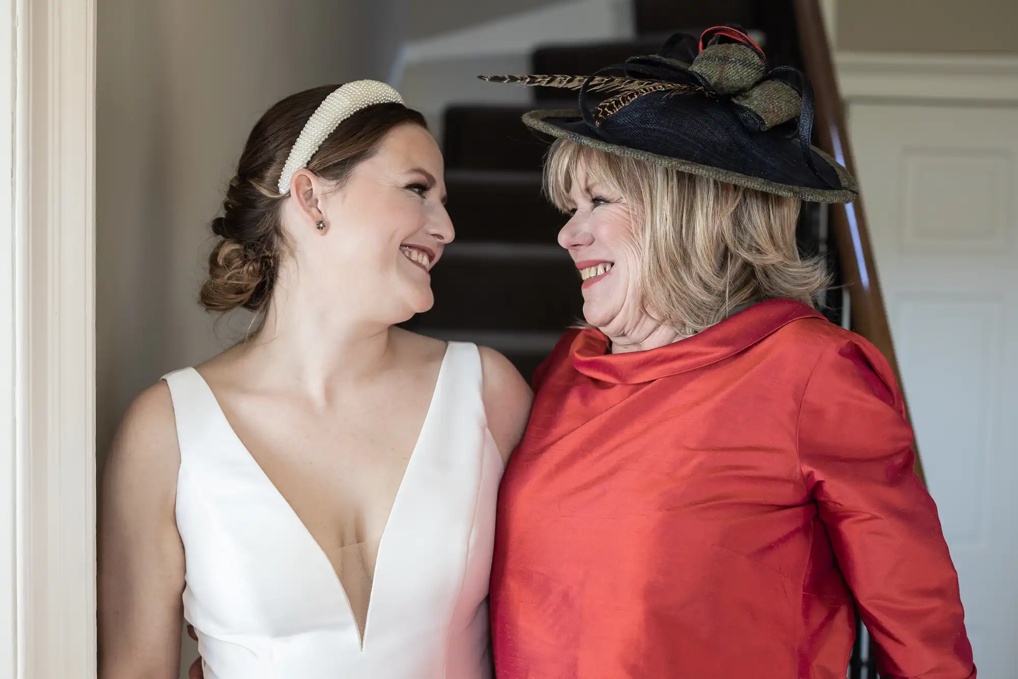 A bride in a white dress and a woman in a red outfit with a decorative hat share a moment of laughter while looking at each other.