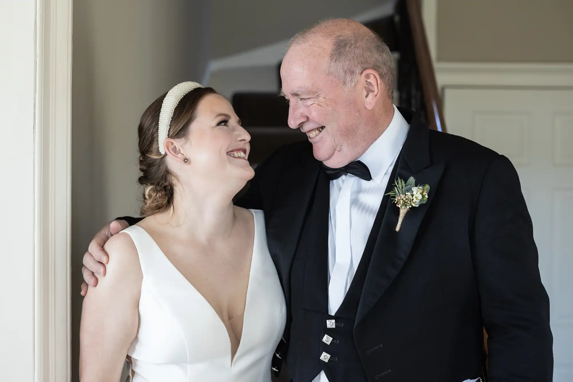 A smiling woman in a white dress stands closely with an elderly man in formal attire, both looking at each other warmly inside a building.