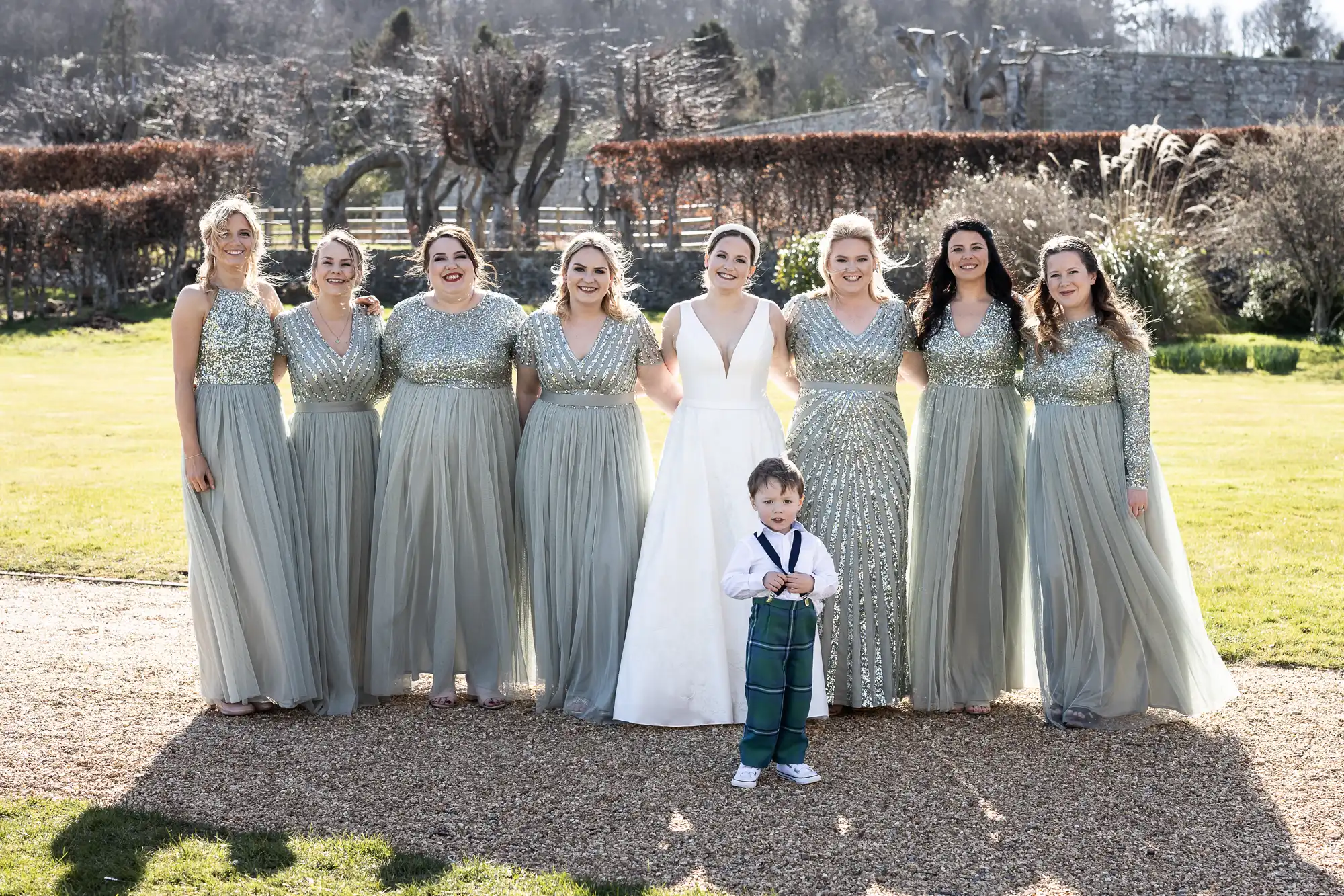 A bride in a white dress stands outside with eight bridesmaids in blue-green dresses, and a young boy in suspenders stands in front of them. They are all posing for a photo on a sunny day.