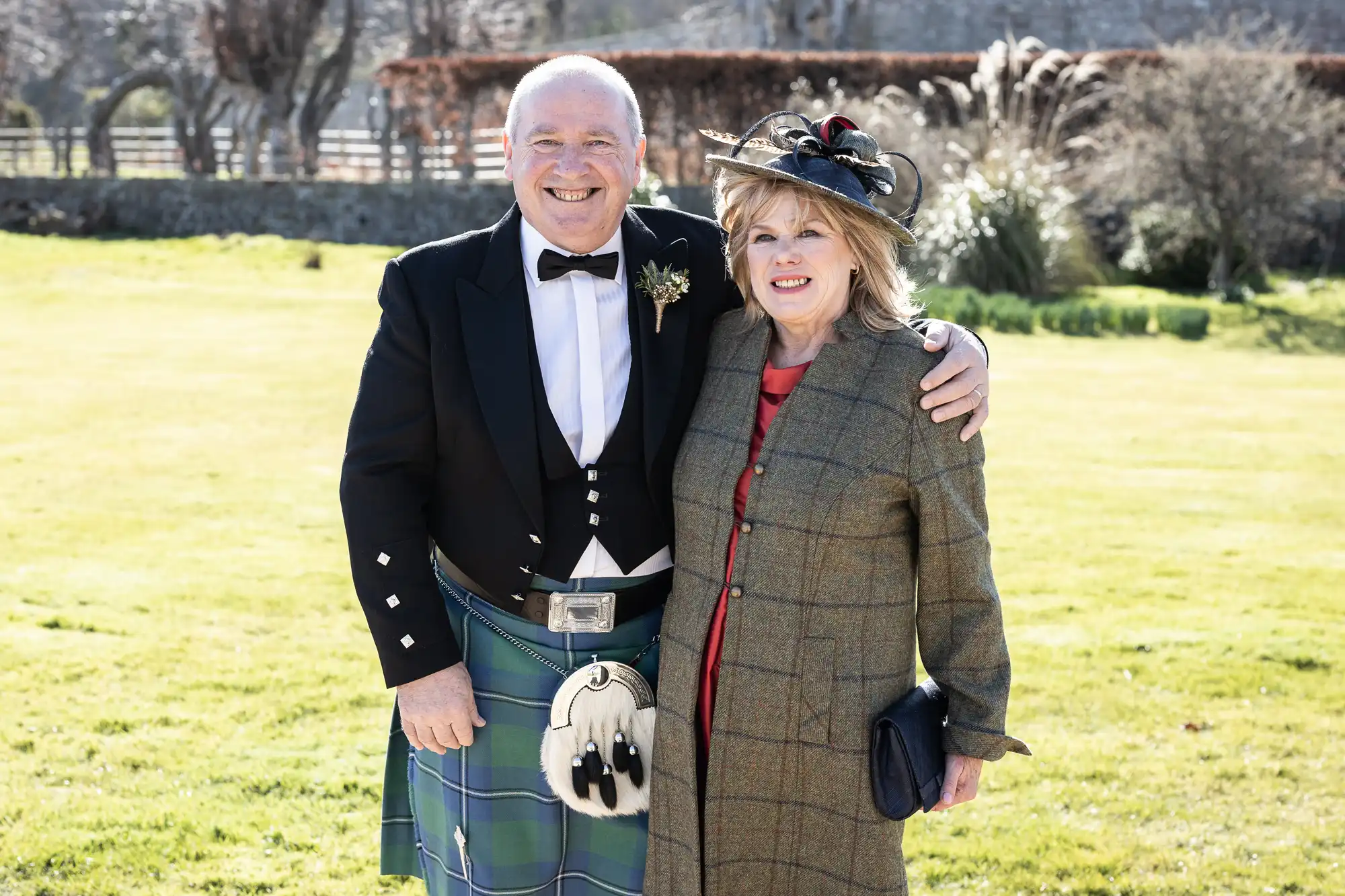 A man in a traditional Scottish kilt and a woman in a plaid coat stand outside on a sunny day, smiling at the camera with arms around each other.