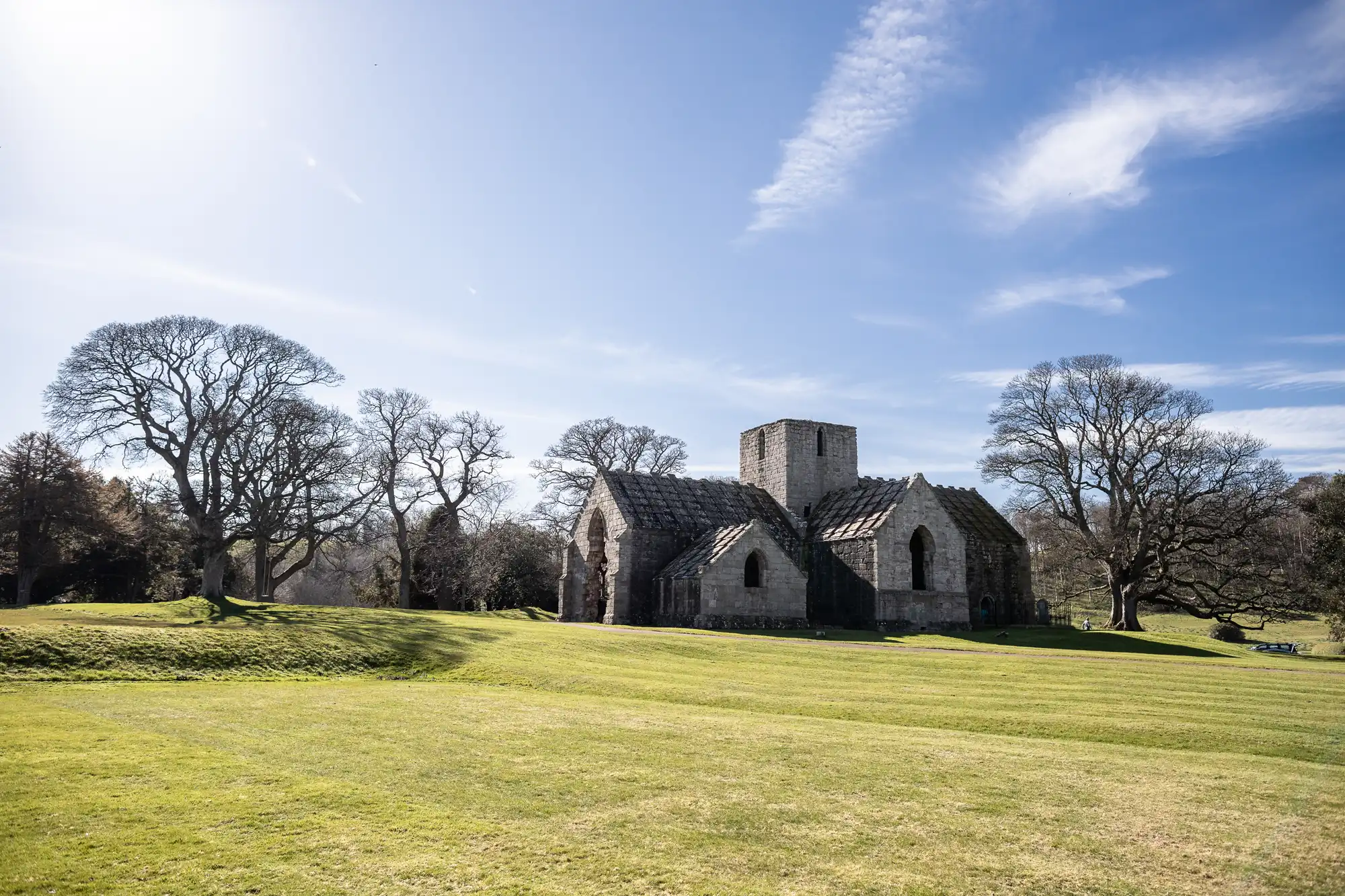 A historic stone church stands in a grassy field under a blue sky with scattered clouds, surrounded by leafless trees.