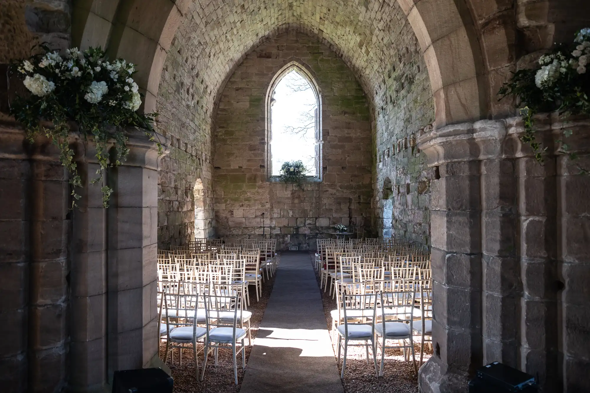 Old stone chapel with arched ceiling, rows of chairs set up for a wedding, and floral arrangements on stone ledges. Bright natural light illuminates the space through a large window at the end.