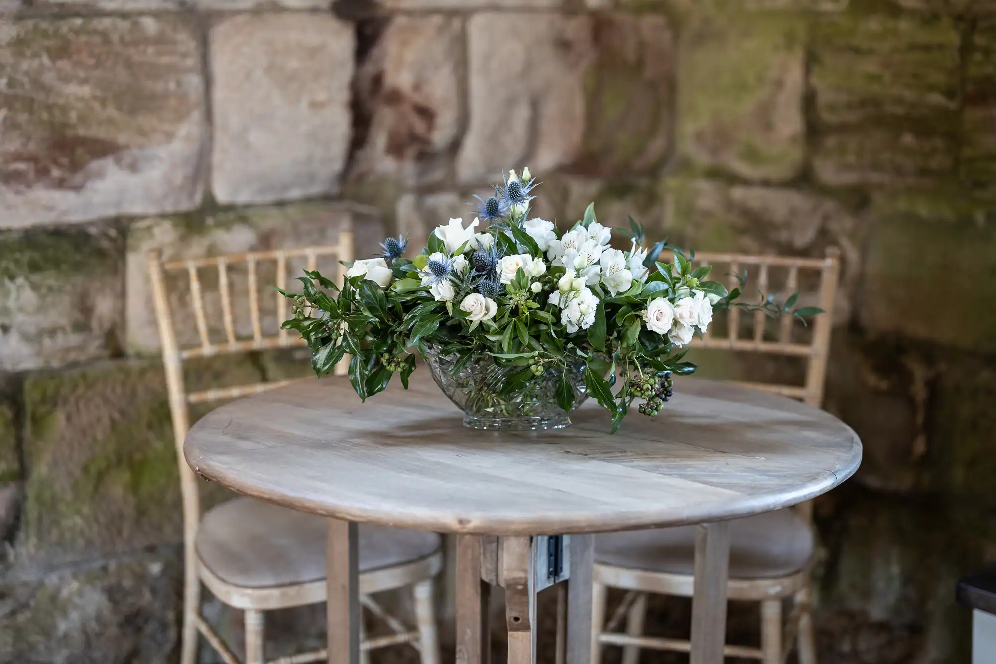 A round wooden table with a floral arrangement of white and blue flowers in a glass vase, placed in front of a stone wall. Two wooden chairs are positioned on either side of the table.