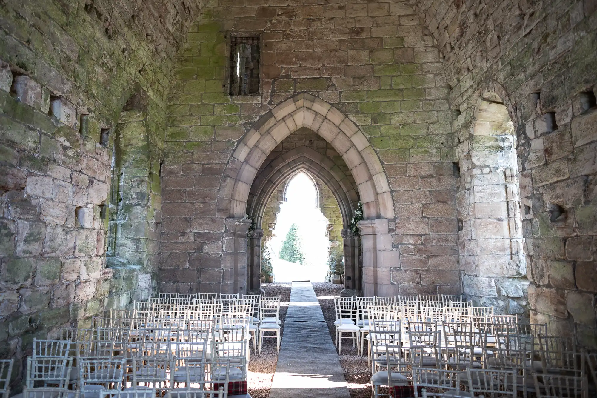 Interior of an old stone church with rows of white chairs arranged in front of a large arched doorway that opens to an outside view. Moss and weathering visible on the stone walls.