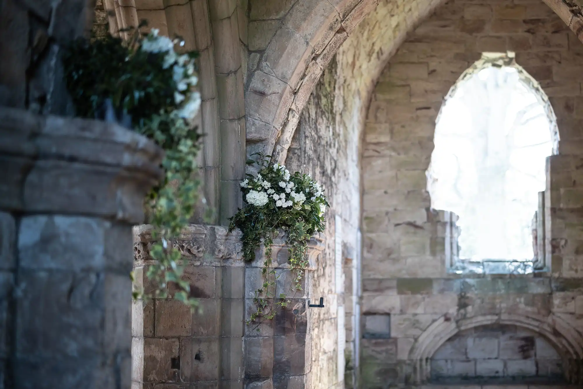 A stone archway adorned with white floral arrangements inside a historic, dilapidated stone building. Sunlight filters through an arched window, illuminating the ancient structure.