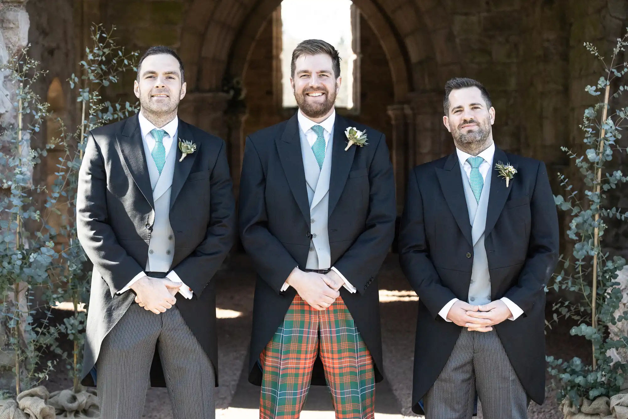 Three men in formal attire stand together, smiling, in front of an arched stone structure. The central figure wears a kilt with plaid patterns, while the others wear matching gray trousers and morning coats.