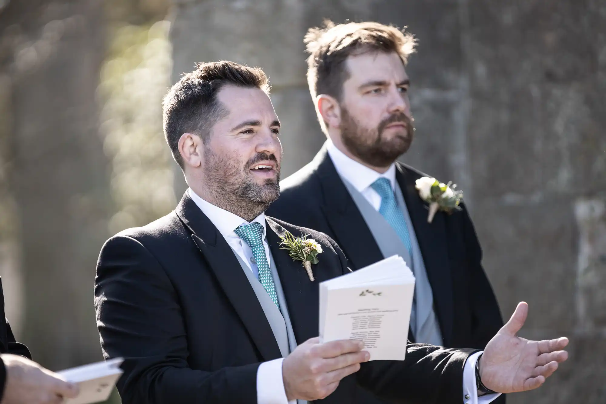 Two men dressed in suits hold open booklets, standing outdoors. Both wear boutonnieres.