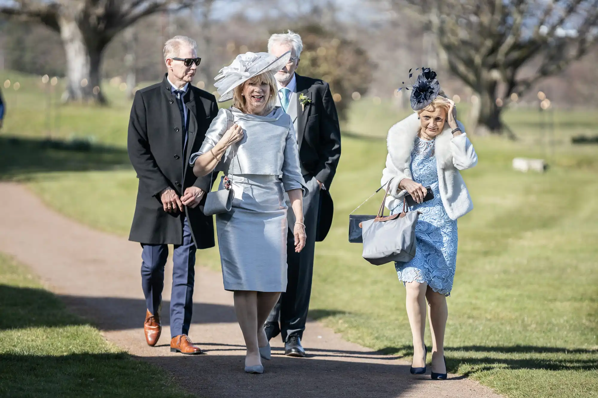 Four people dressed up for an outdoor event are walking together on a path. The two women are wearing hats and formal dresses, while the two men are in suits. Trees and grass are visible in the background.