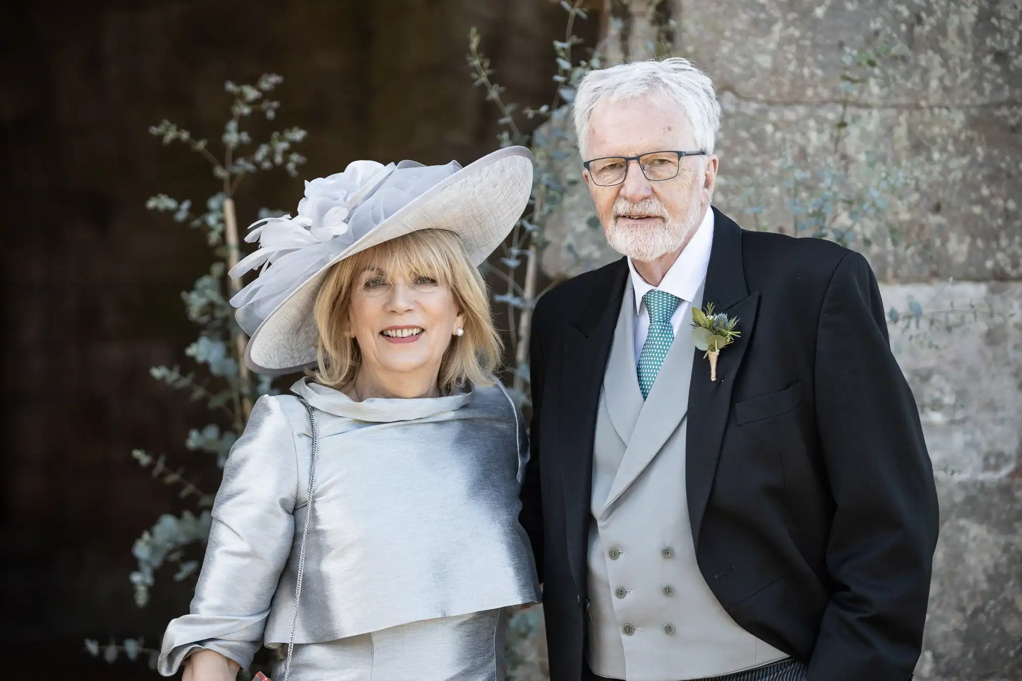 An elderly couple is posing together outdoors, with the woman in a silver dress and large hat and the man in a suit with a boutonnière.