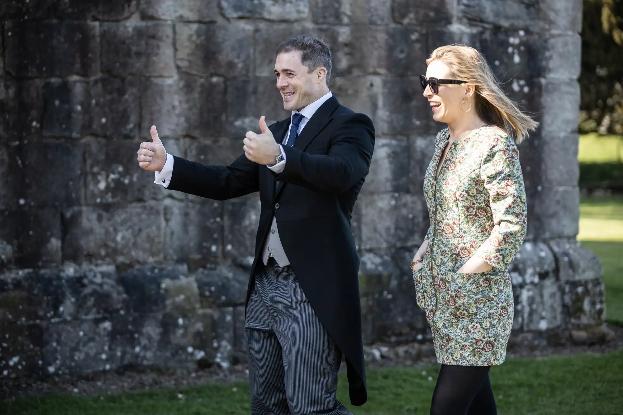 A man in a dark suit and a woman in a floral dress walk outside, both smiling. The man is giving two thumbs up. They are in front of a stone wall.