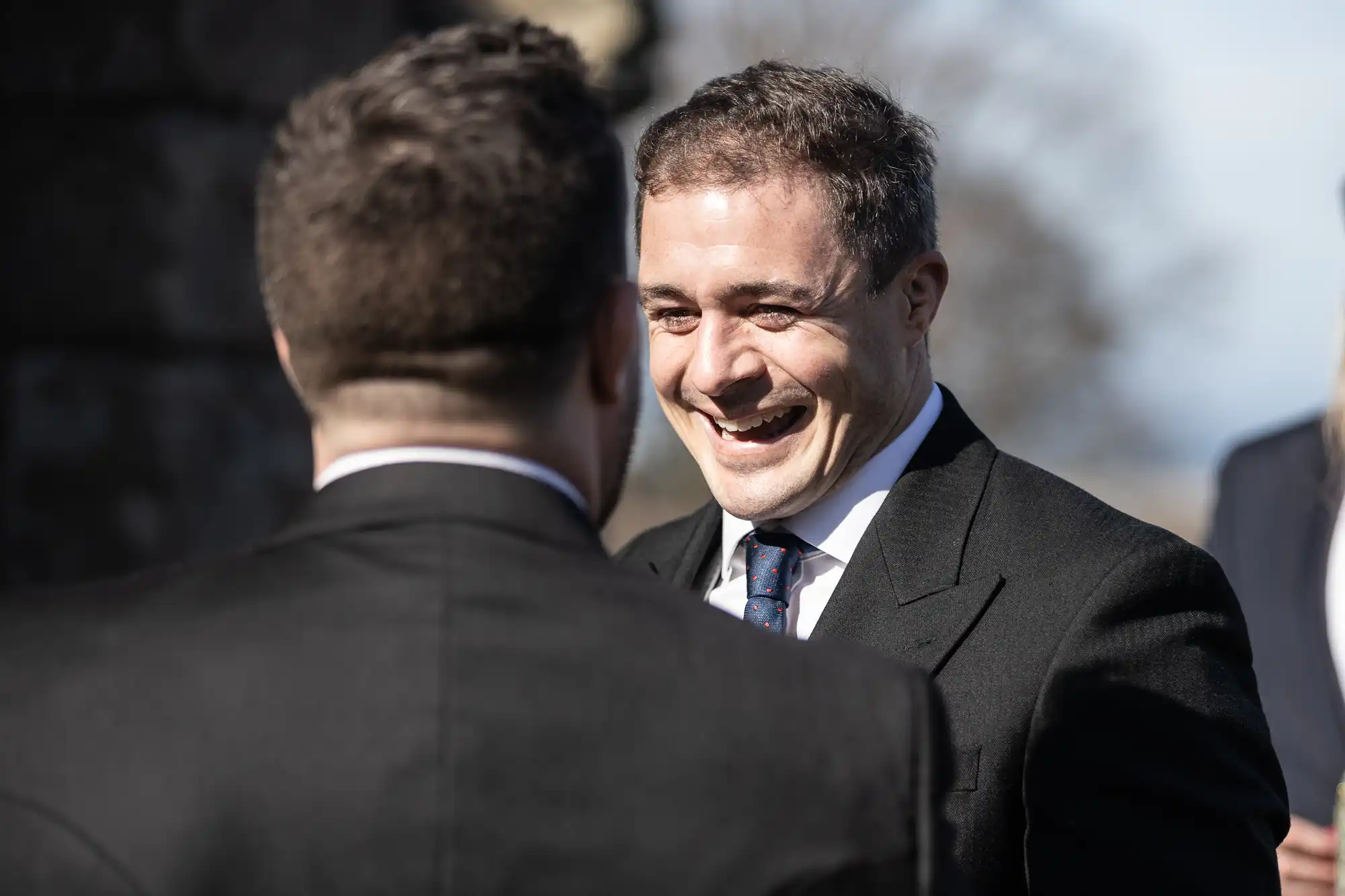 Two men in dark suits engage in a conversation outdoors; the man facing the camera is smiling broadly.