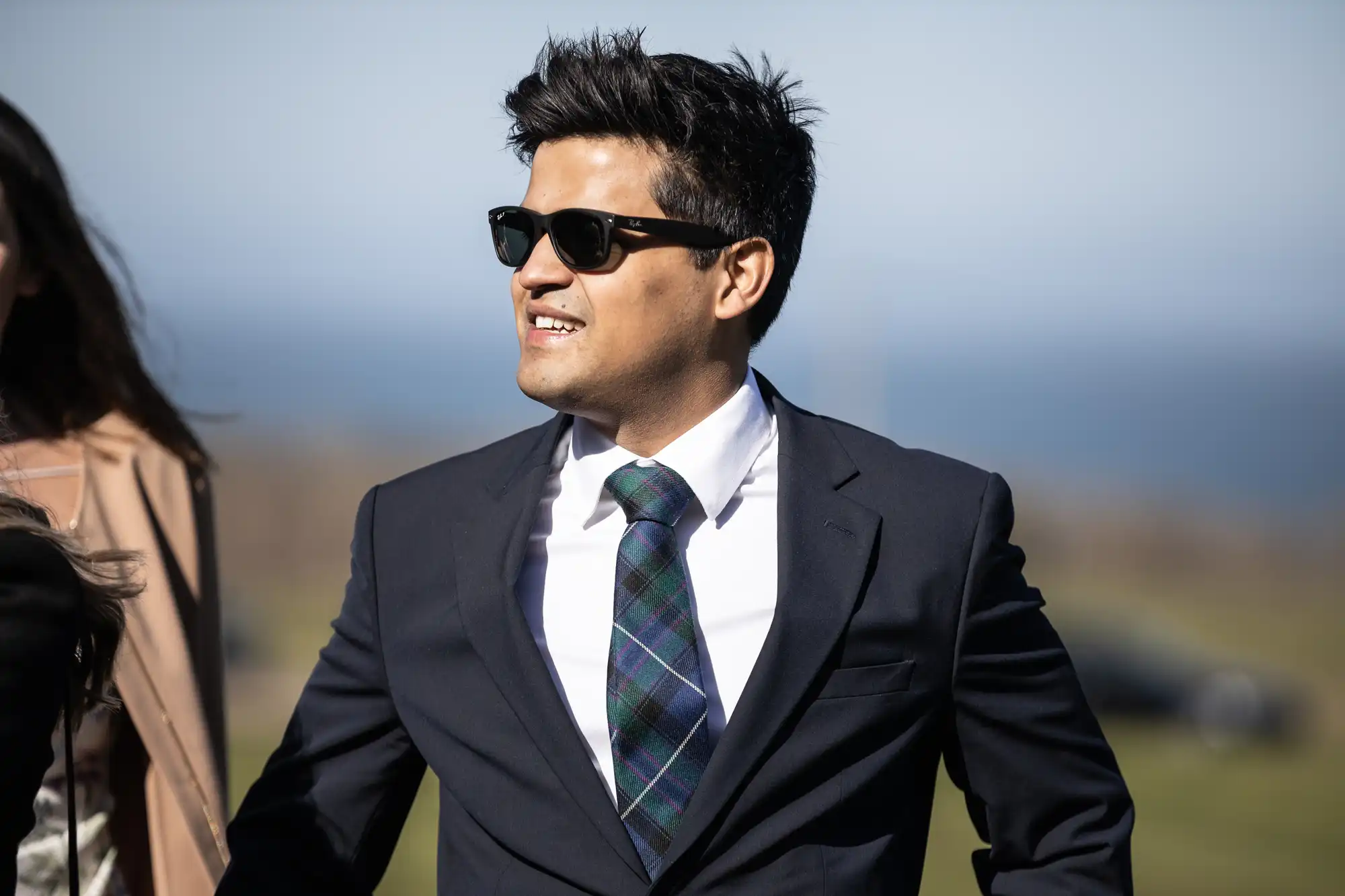 A man in a suit and tie, wearing sunglasses, stands outdoors with a faint background of grass and sky.