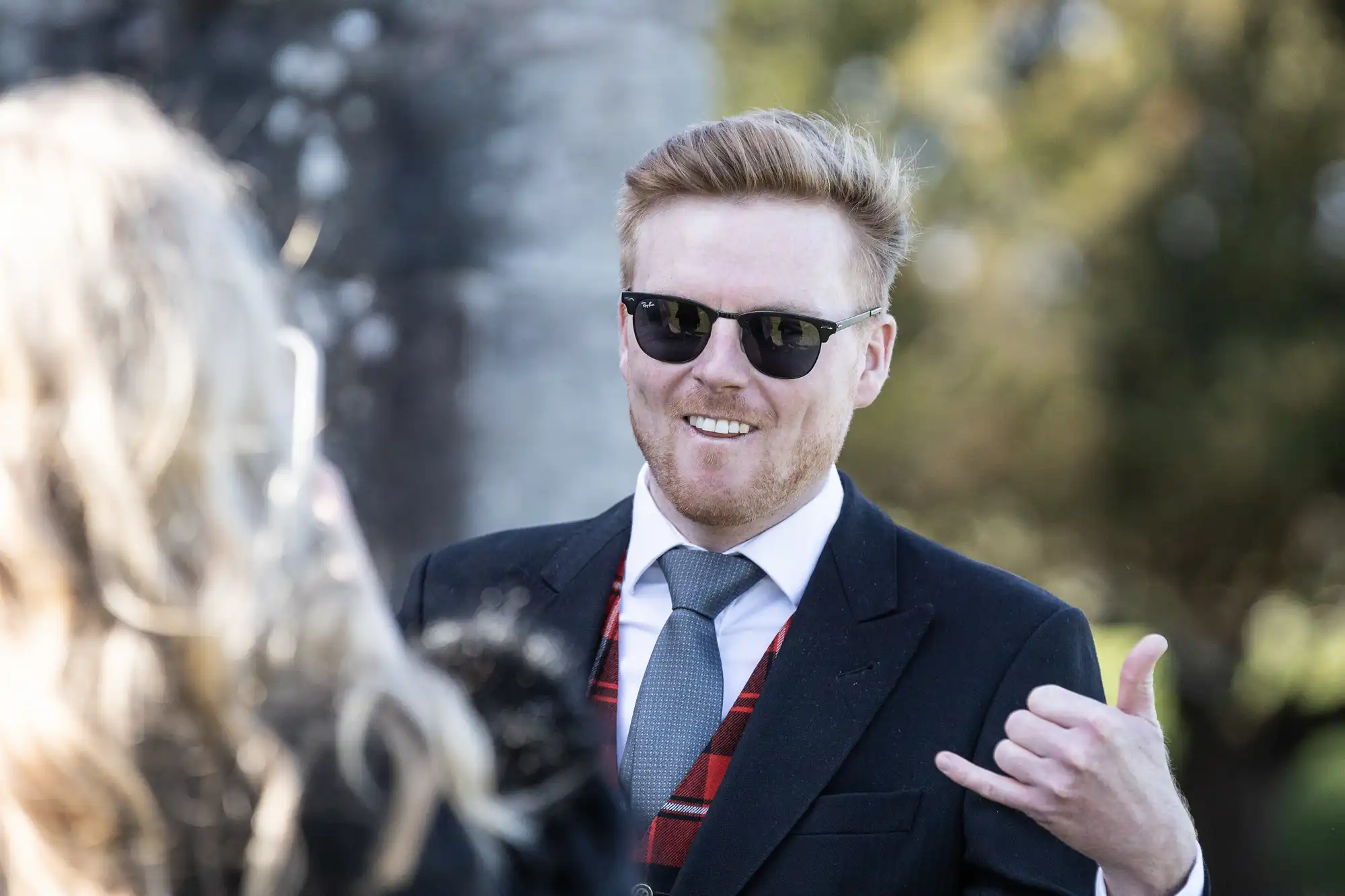 Man in a suit and tie wearing sunglasses gesturing with his right hand while talking to a person with long blonde hair in an outdoor setting.