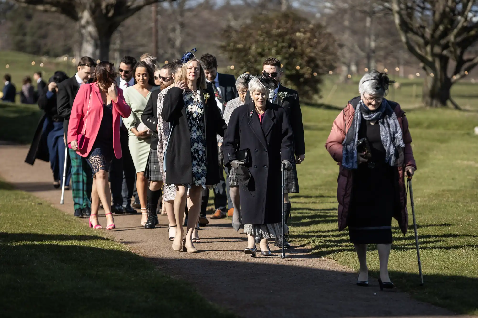 A group of people, dressed formally, walk along a pathway in a park. Two older individuals are using walking sticks, leading the group. Trees and grassy areas are visible in the background.