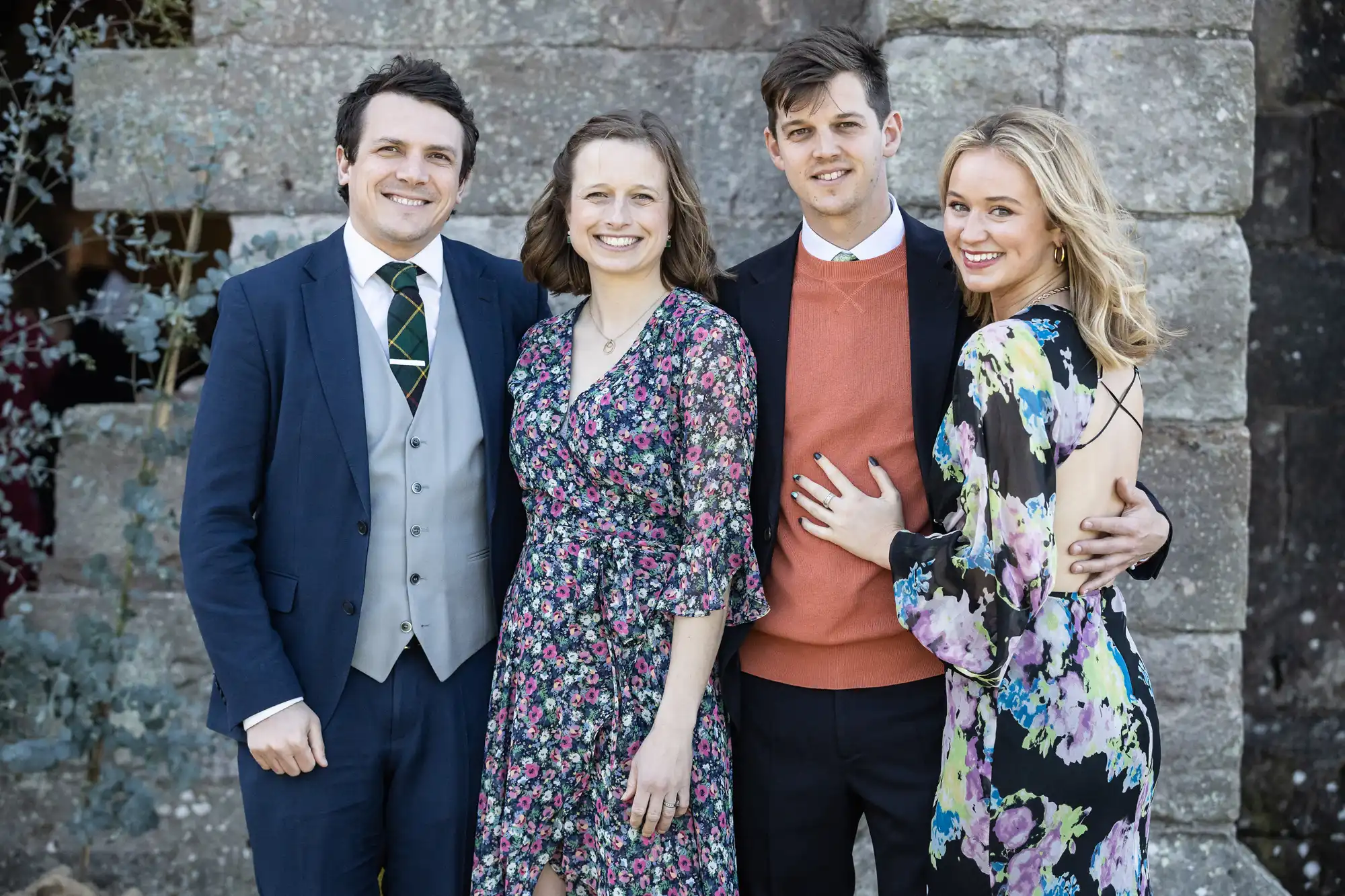 Four people, two men and two women, stand closely together in front of a stone wall, smiling. The individuals are dressed in formal attire, with the women wearing floral dresses and the men in suits.