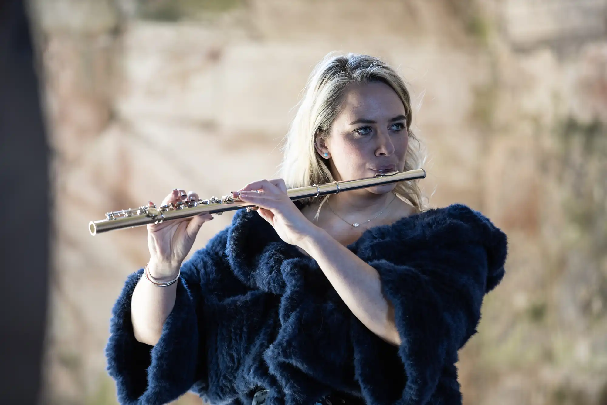 A woman in a dark, textured top plays a flute outdoors with a blurred, earthy background.