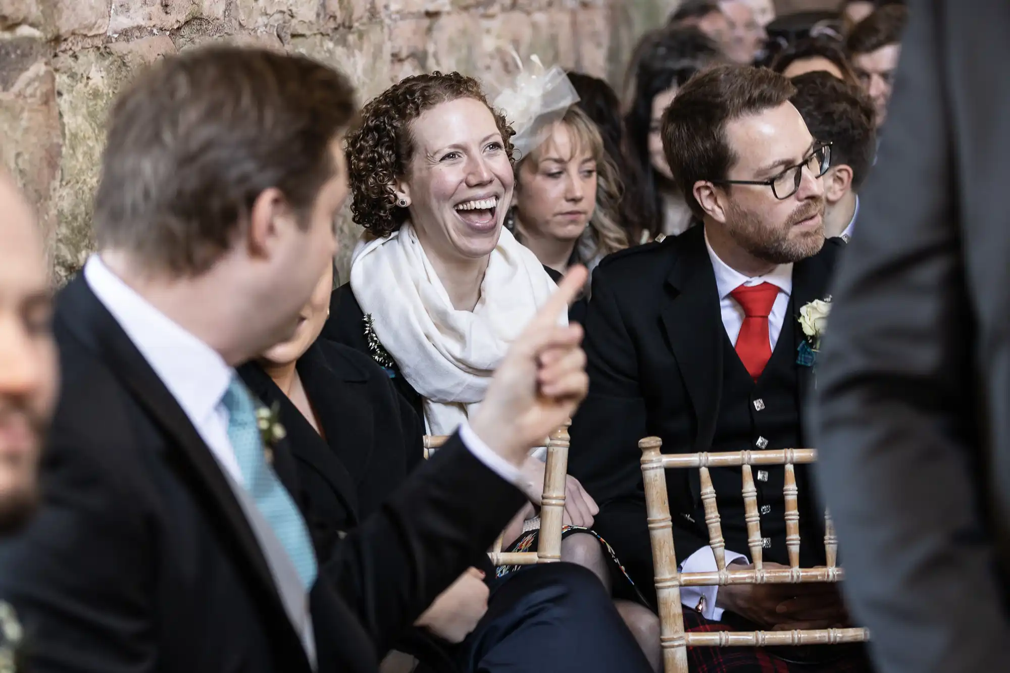 Several people are seated and engaged in conversation at an indoor event. A woman with a wide smile stands out among them. The background features a rustic brick wall.