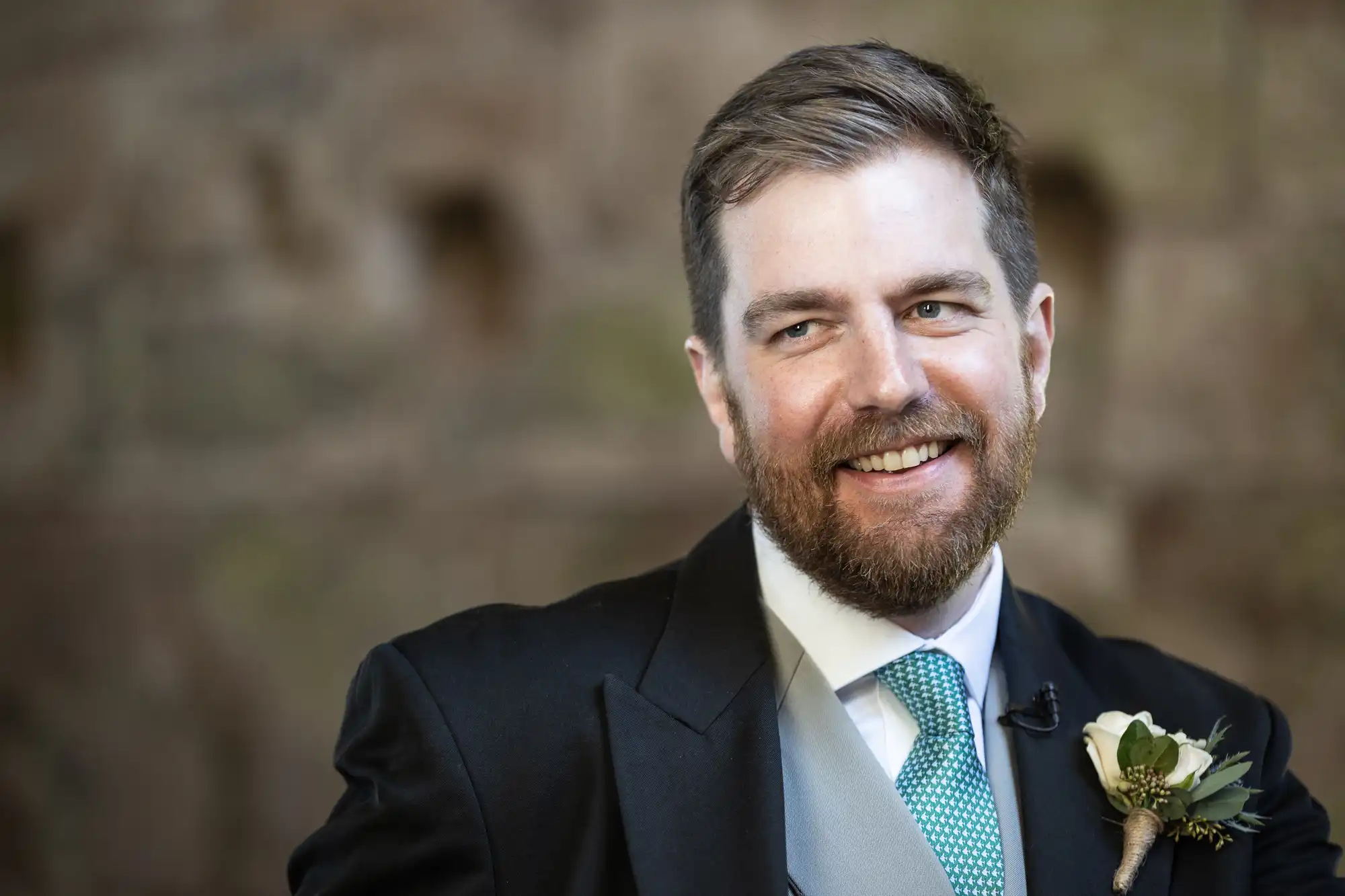 A man with a beard and short hair wearing a black suit, light blue tie, and a boutonniere smiles while looking to the side. The background is blurred.