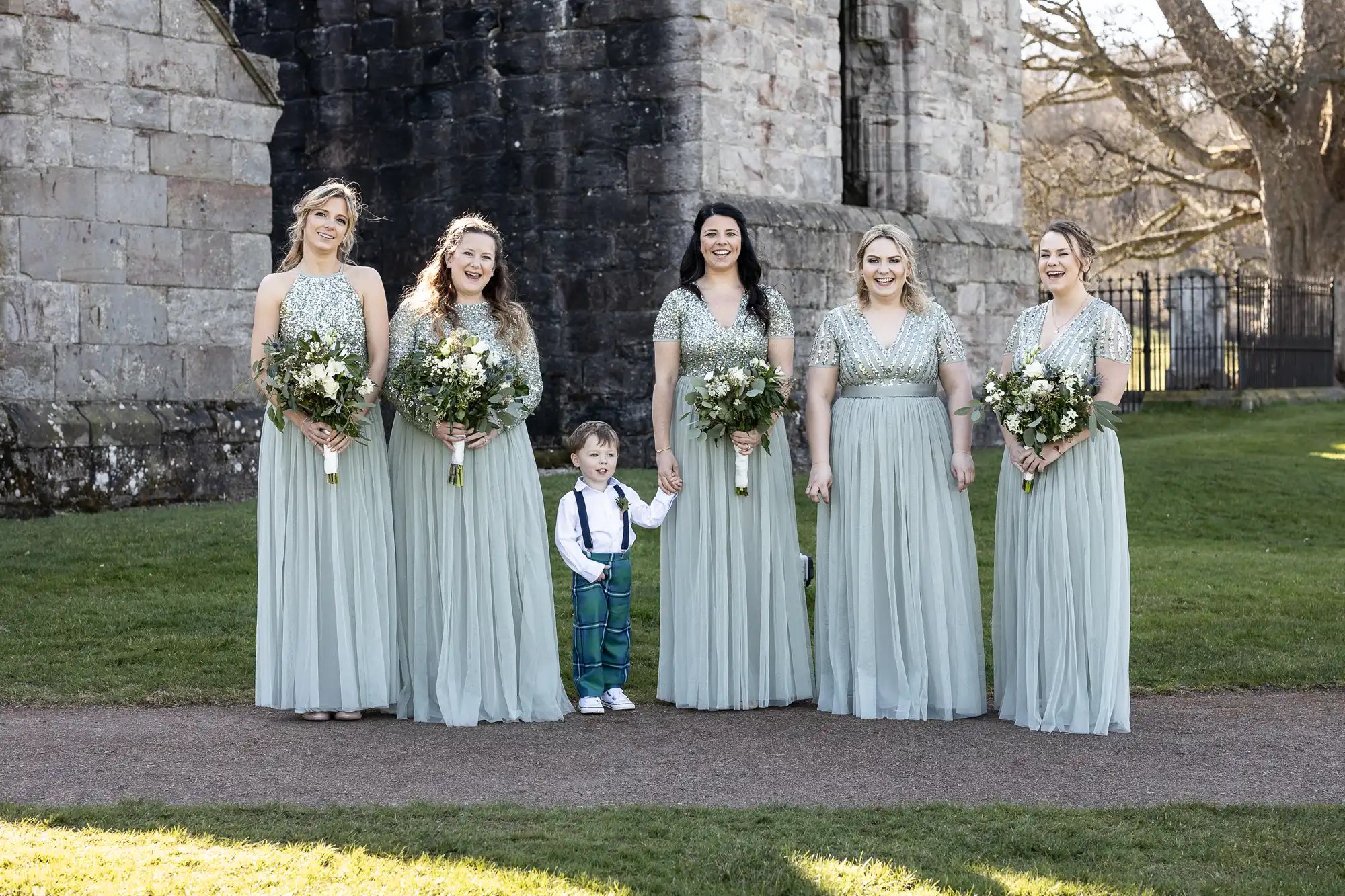 Five women in light green dresses stand outdoors next to a small boy in a white shirt and blue suspenders, all holding floral bouquets. A stone building and grassy area are in the background.