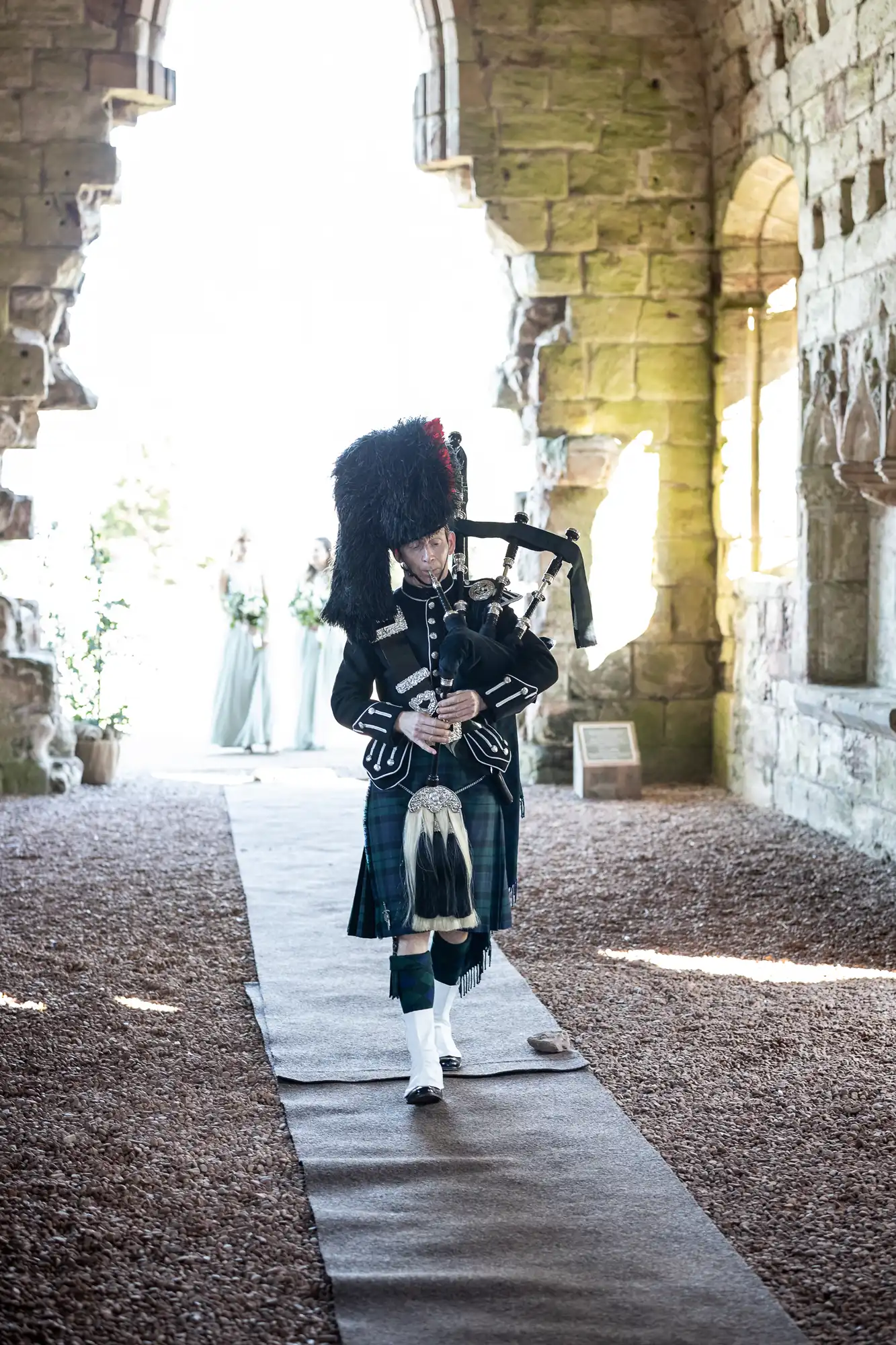 A person in traditional Scottish attire plays bagpipes while walking on a carpeted path through a stone archway.