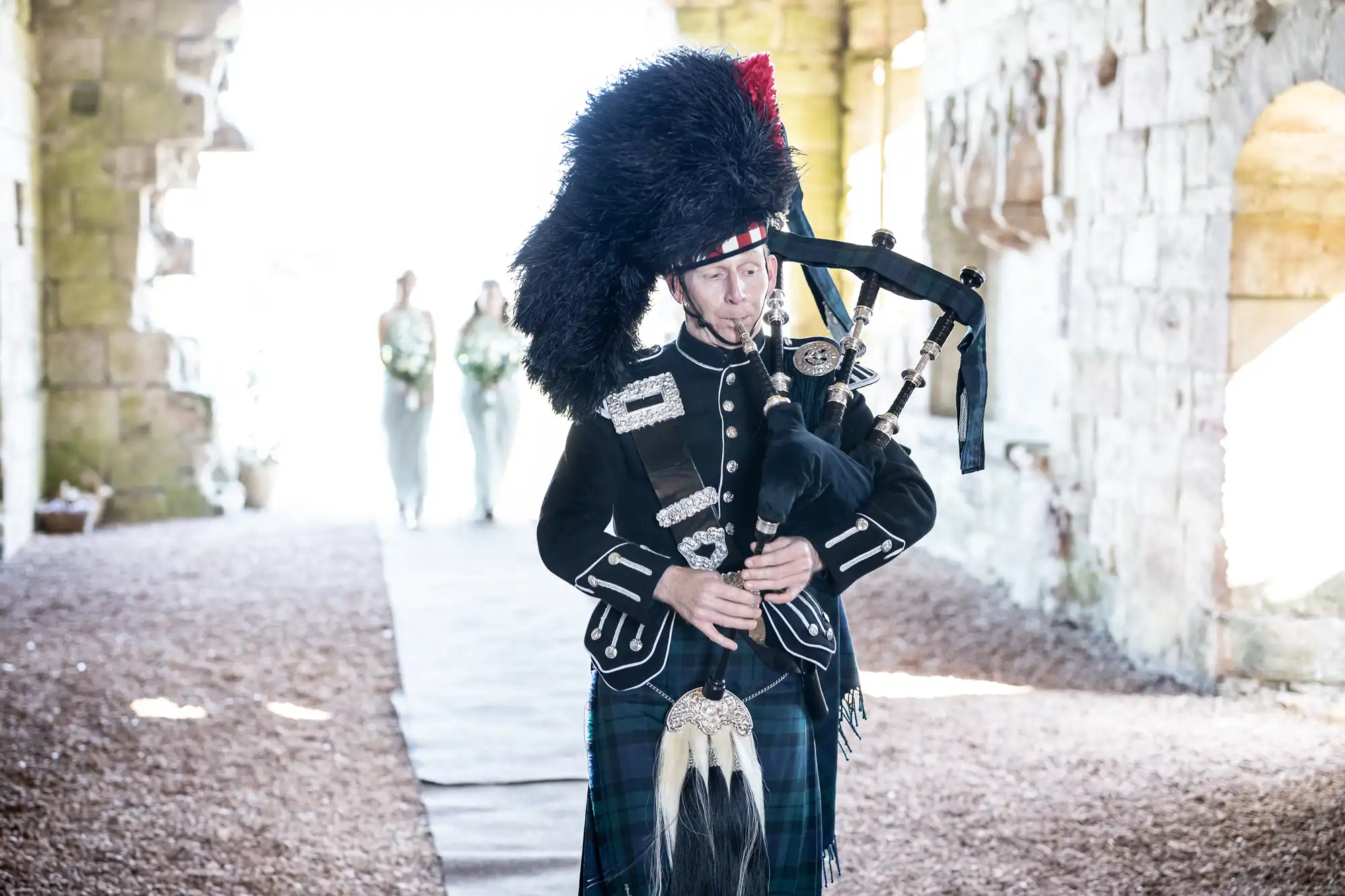 A person dressed in traditional Scottish attire plays bagpipes in an outdoor setting with two individuals in light dresses in the background.