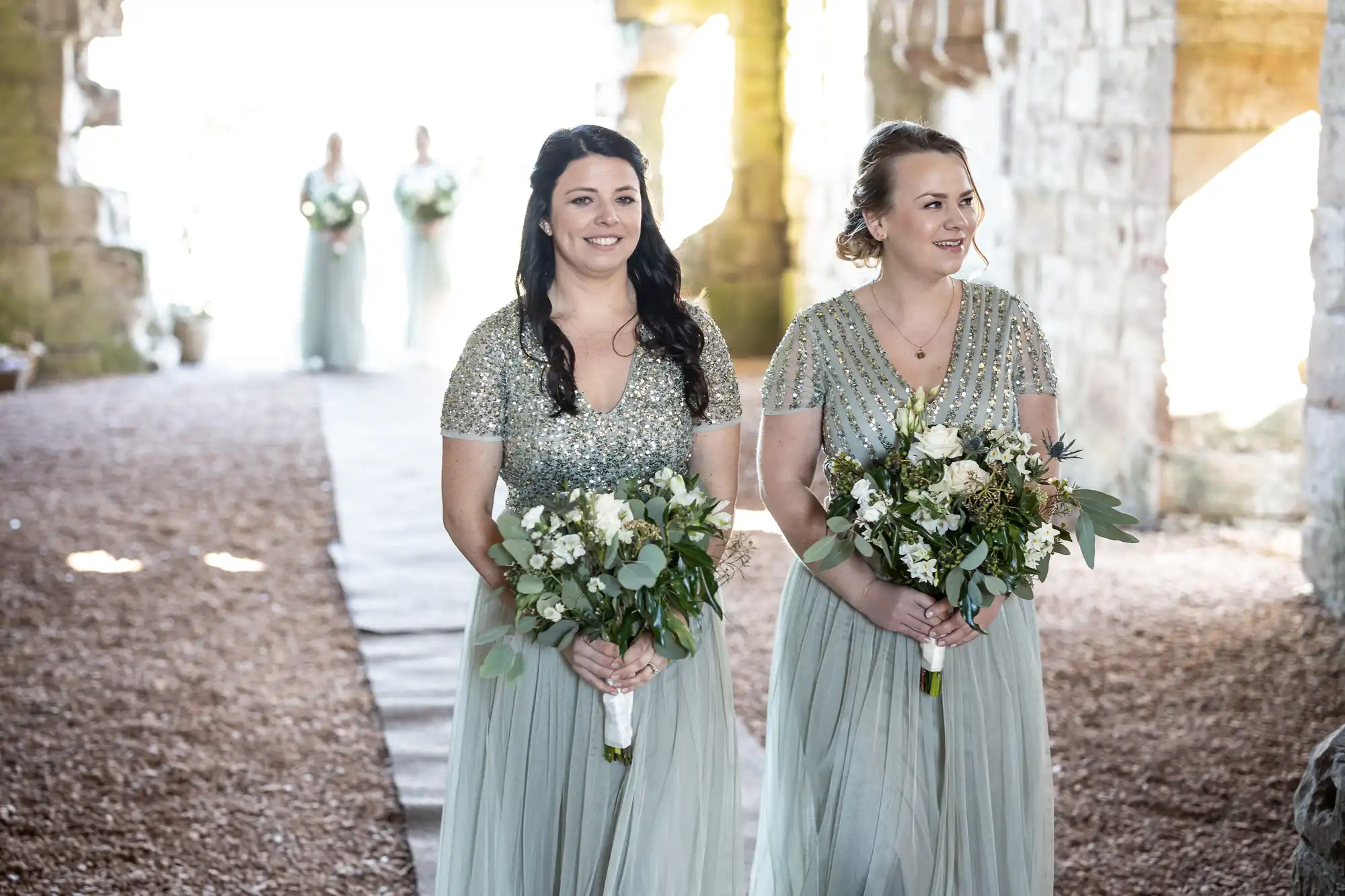 Two bridesmaids in silver sequin gowns holding white and green bouquets walk down an aisle in an outdoor stone venue, with two more bridesmaids in the background.