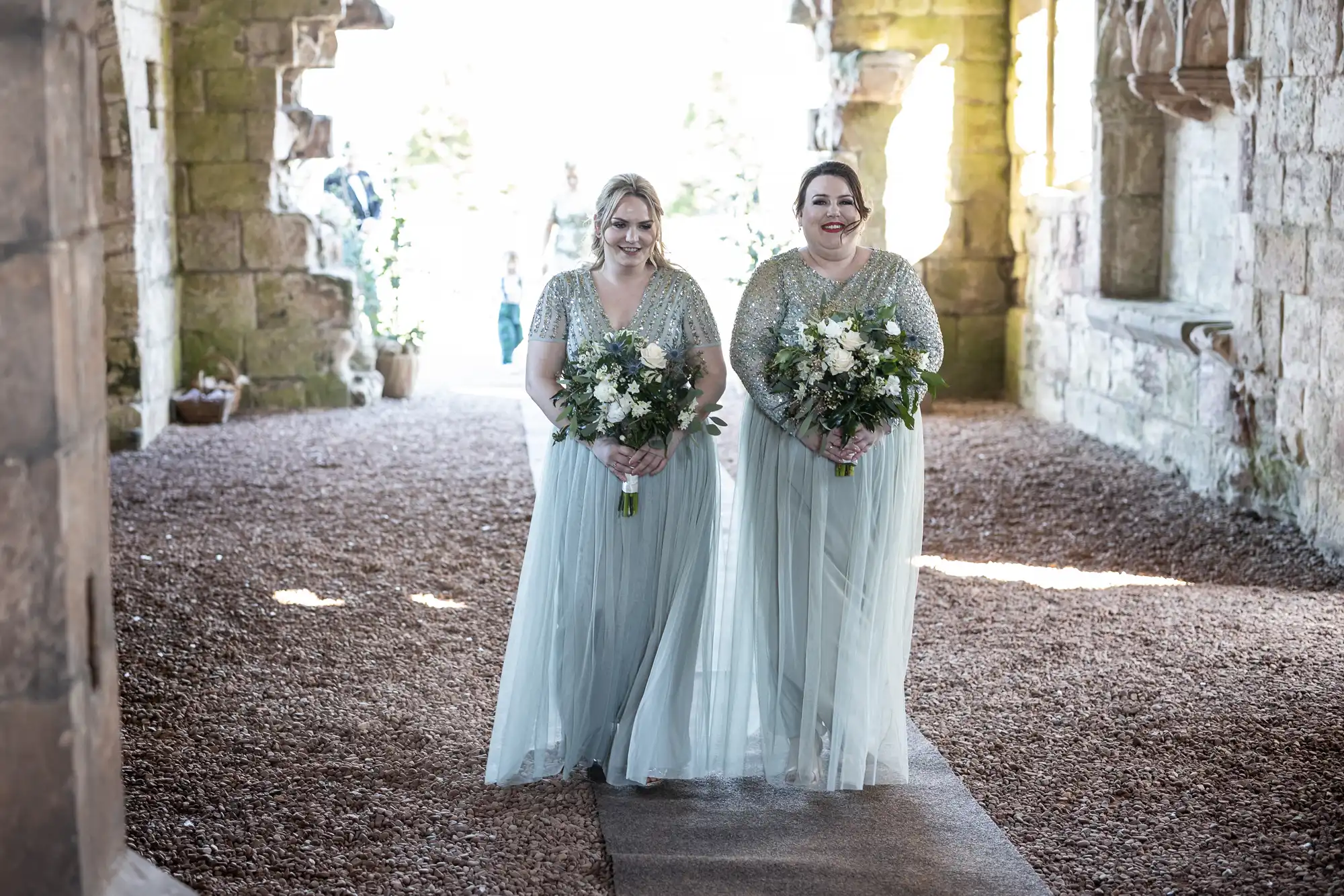 Two bridesmaids in light grey dresses hold bouquets and walk down a gravel path, surrounded by ancient stone walls.