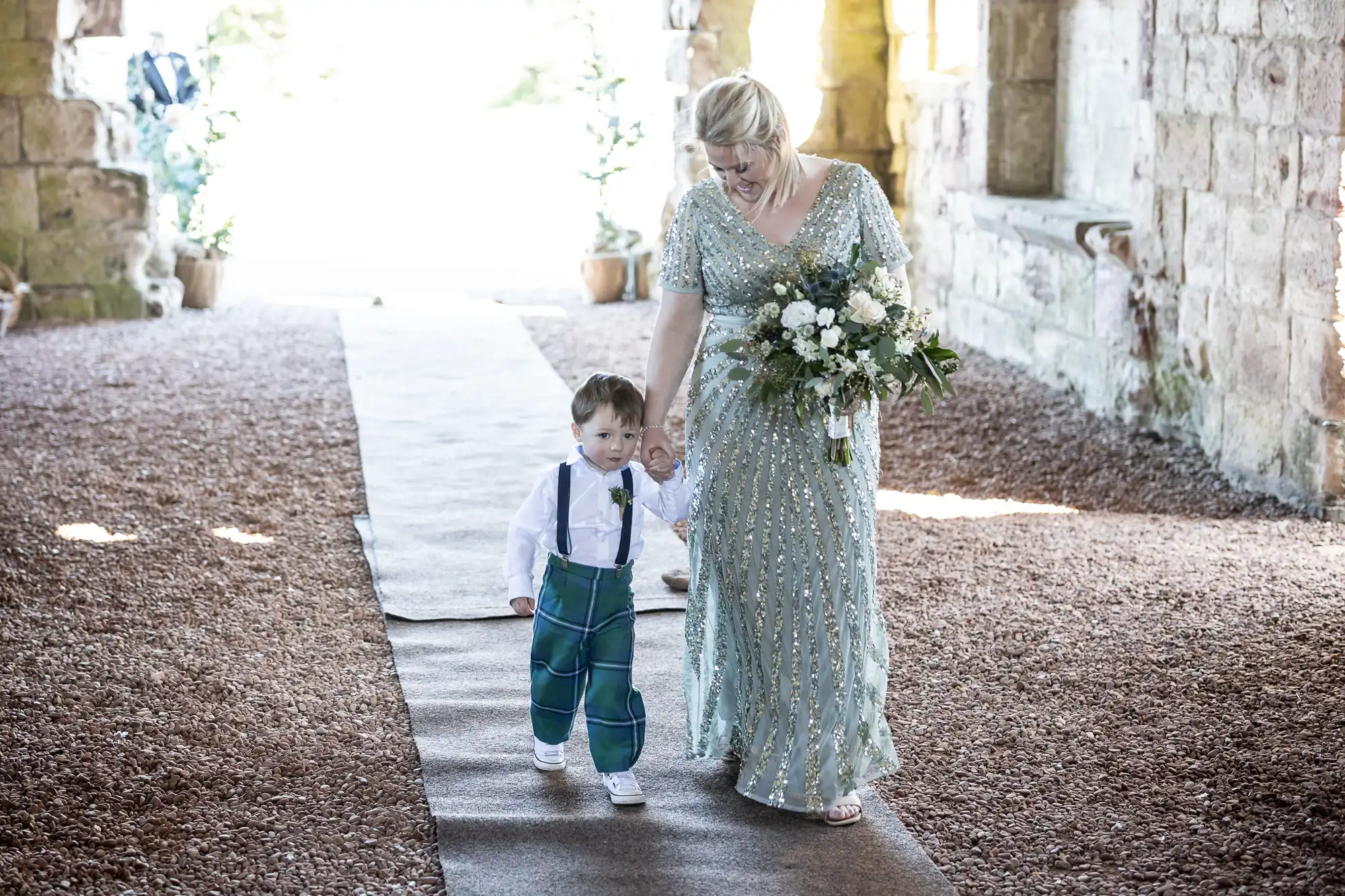 A woman wearing a sparkling dress walks down a stone path holding hands with a young child in suspenders and plaid pants. She carries a bouquet of flowers in her other hand.