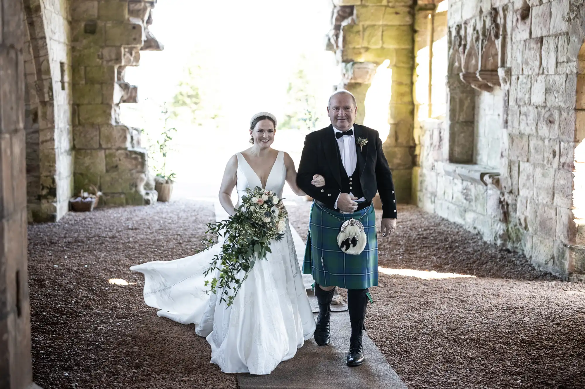 A bride in a white gown and a man in a kilt walk arm in arm down a stone corridor, both smiling. The bride holds a bouquet of flowers and greenery.