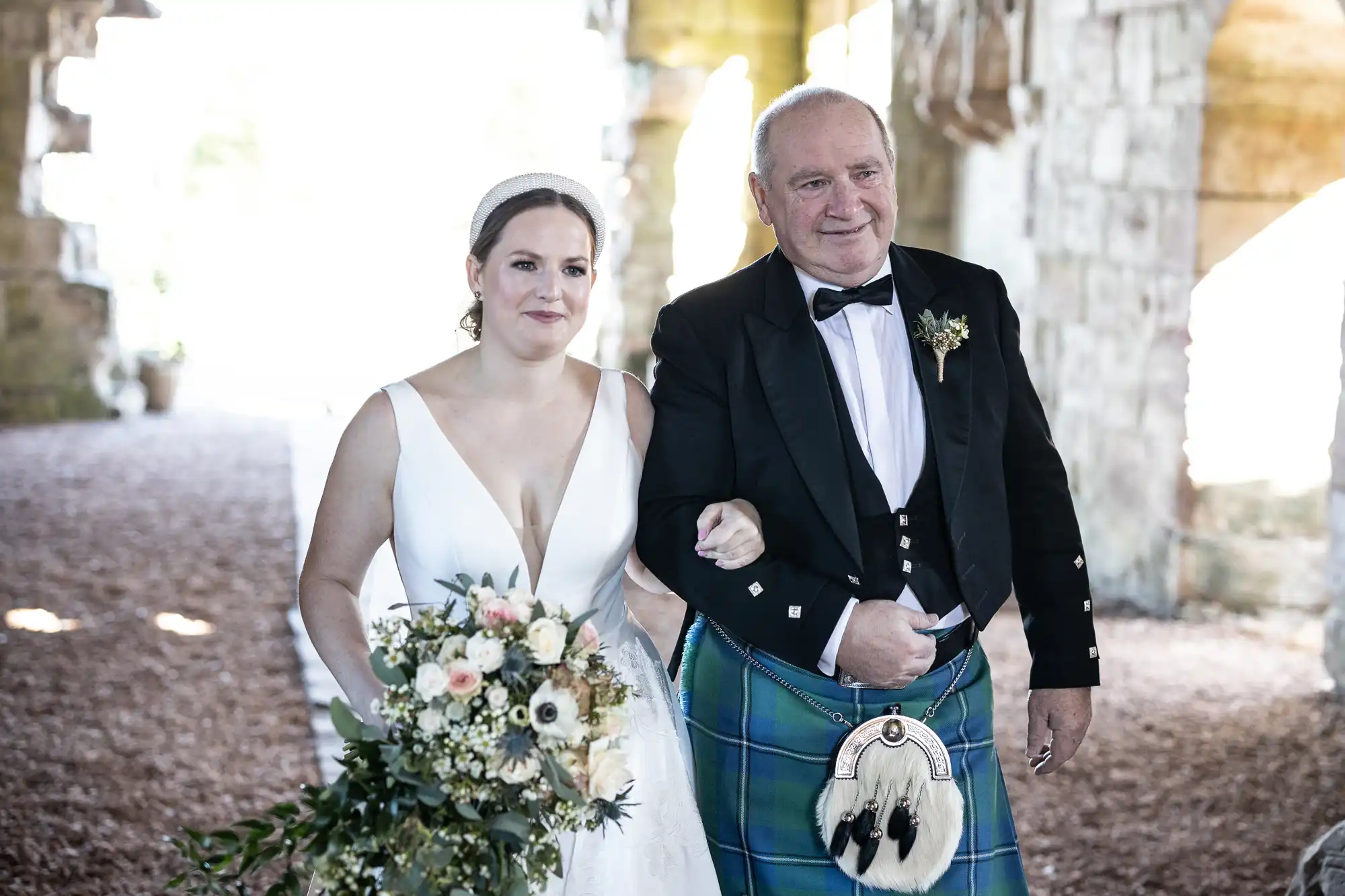 A bride in a white gown and a man in a traditional kilt, likely her father, walk down the aisle at an outdoor wedding ceremony. She holds a bouquet of flowers, and they both smile.