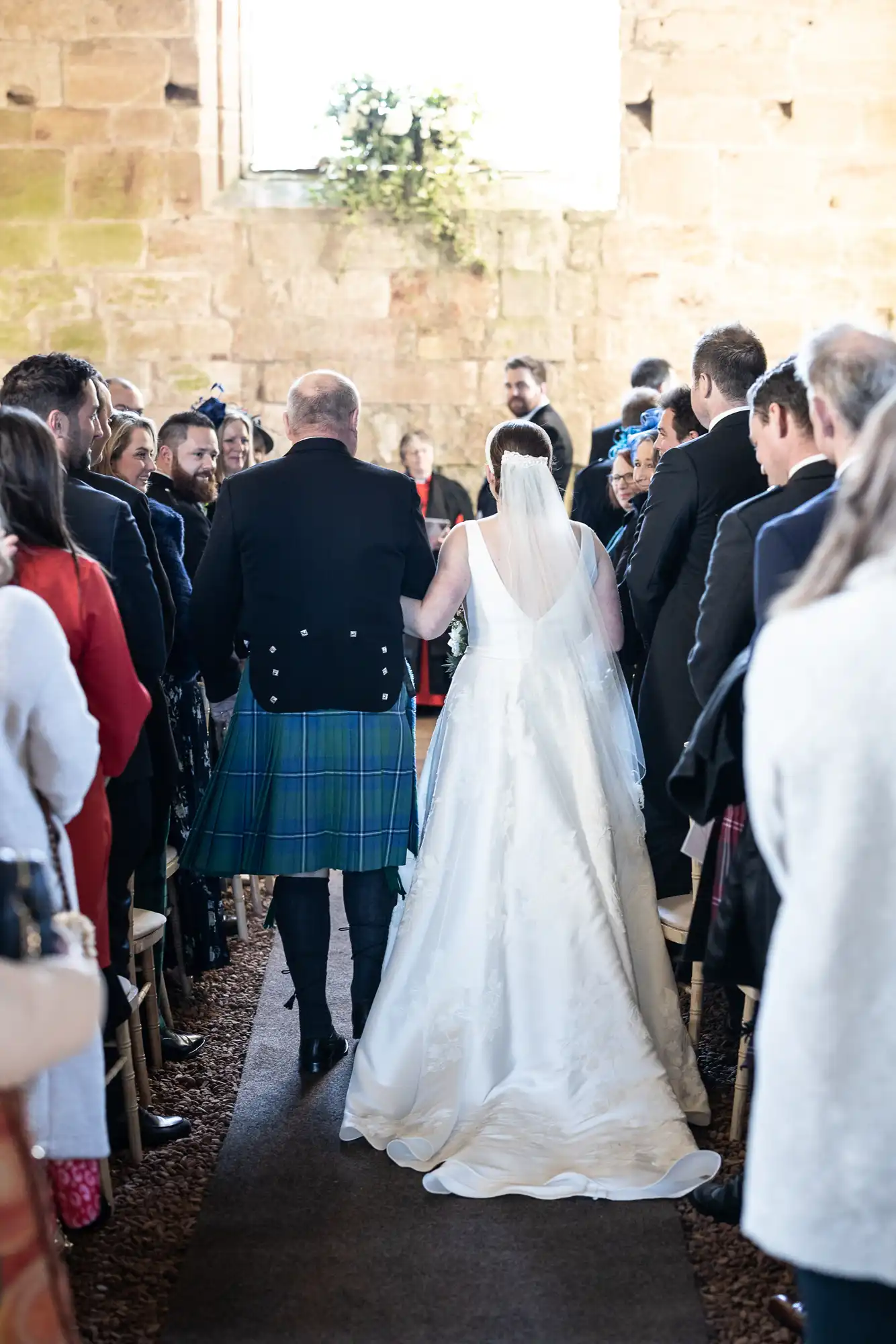 A bride in a white dress walks down the aisle with an older man in a kilt, surrounded by people seated on either side in a stone-walled venue.