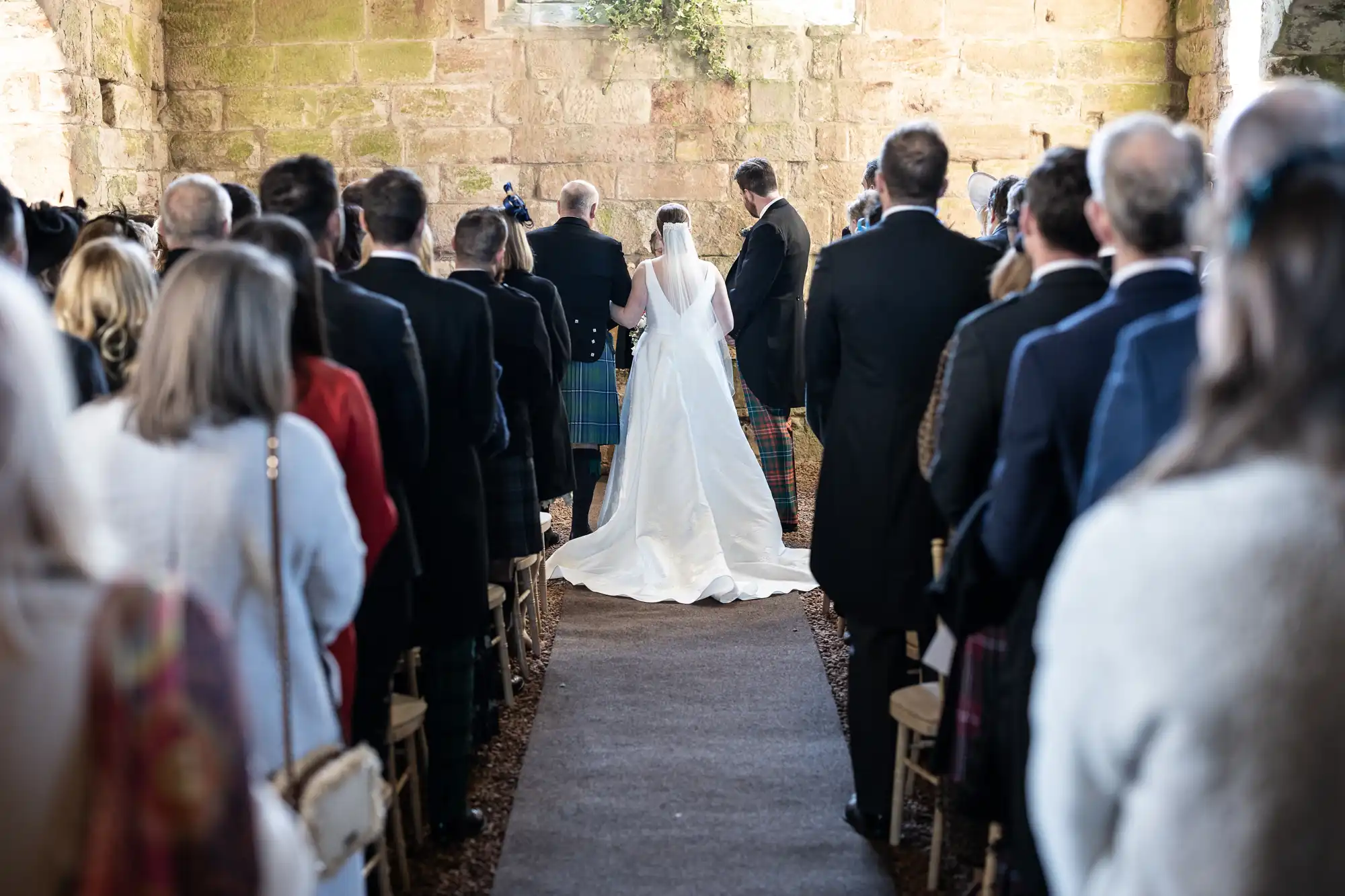 A bride, groom, and officiant stand at the front of an aisle lined with seated wedding guests, captured from the back of the venue. The ceremony is set in a rustic stone building.