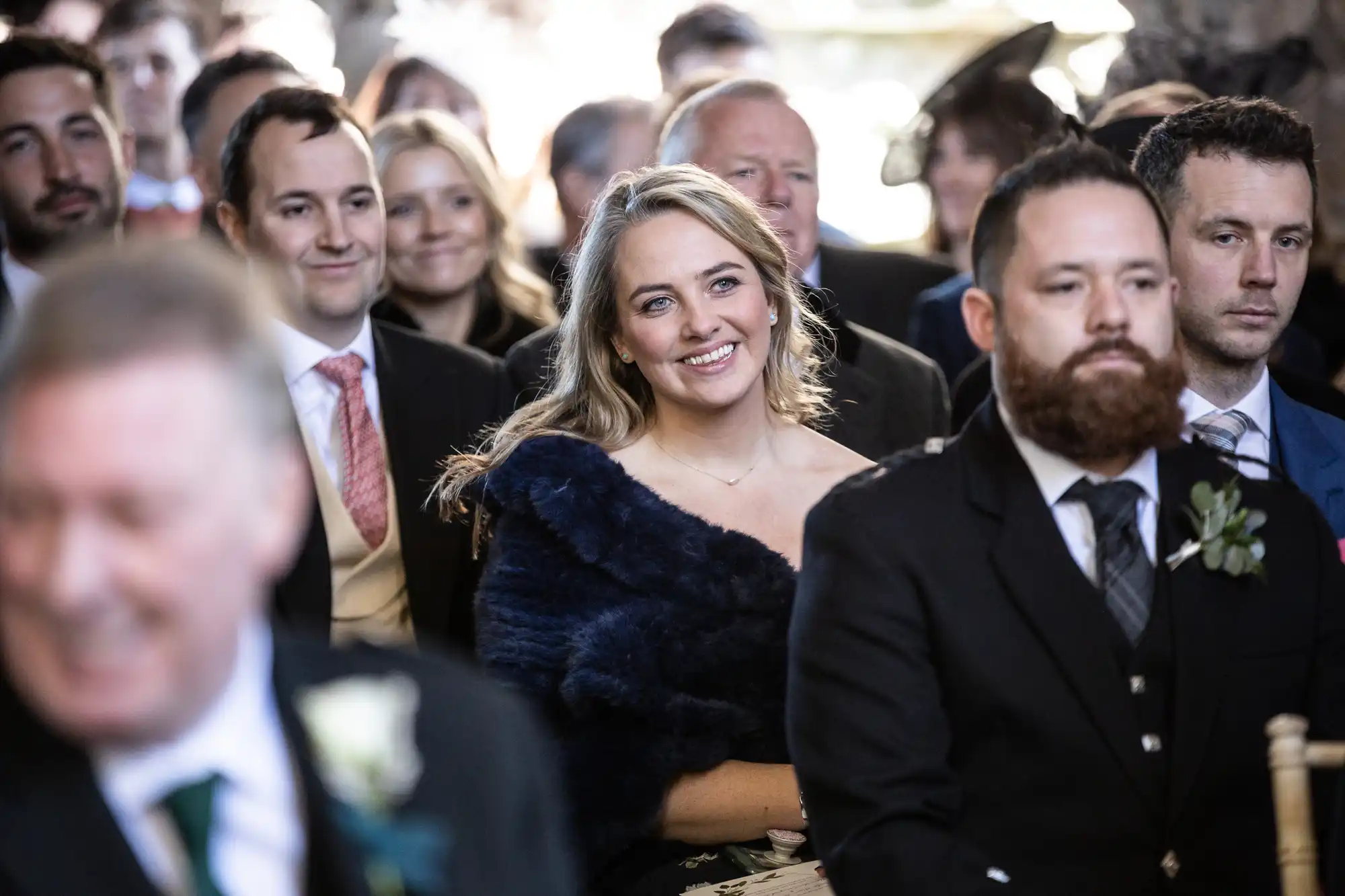 A group of people in formal attire sit indoors at an event, with a woman in the center smiling, draped in a dark shawl.