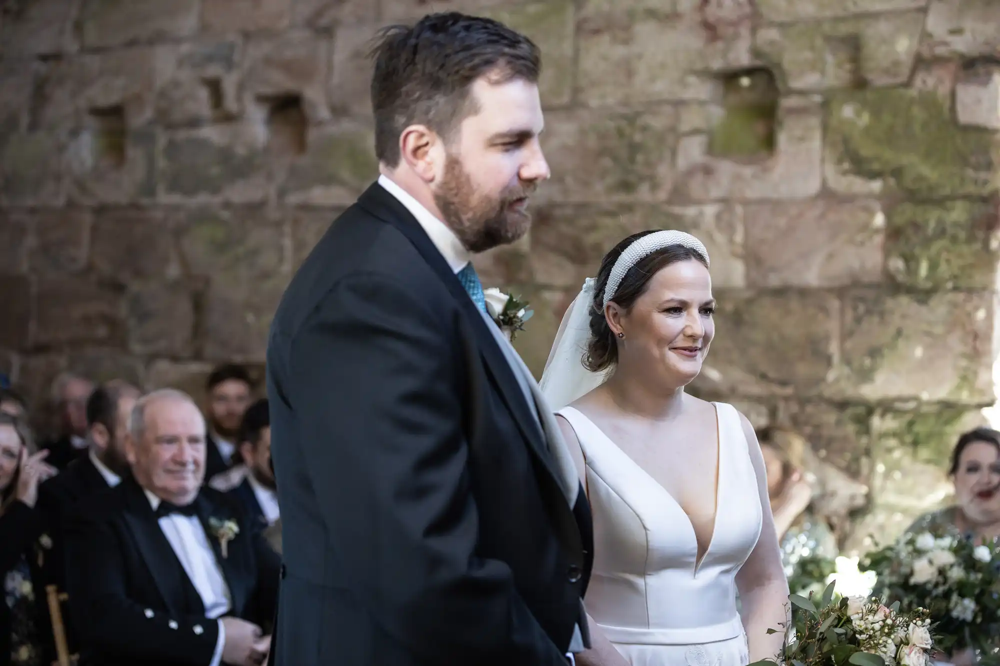 A couple stands together during their wedding ceremony, with the groom wearing a suit and the bride in a white dress and veil. Guests are seated in the background within a rustic stone venue.