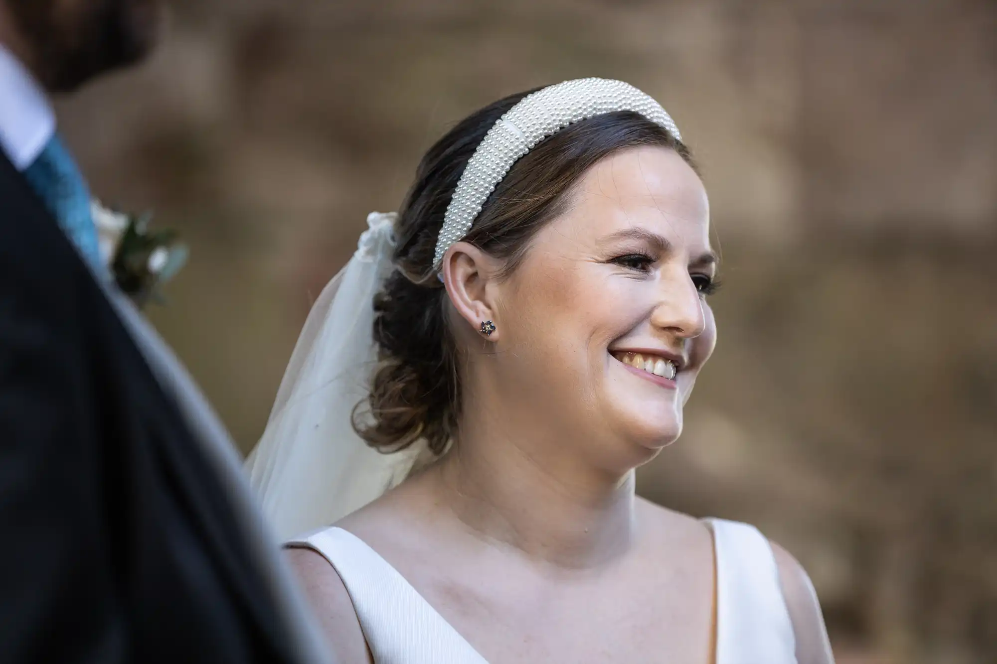 A woman in a white dress and headband smiles while looking to the right. She has a veil attached to the headband. The background is blurred.