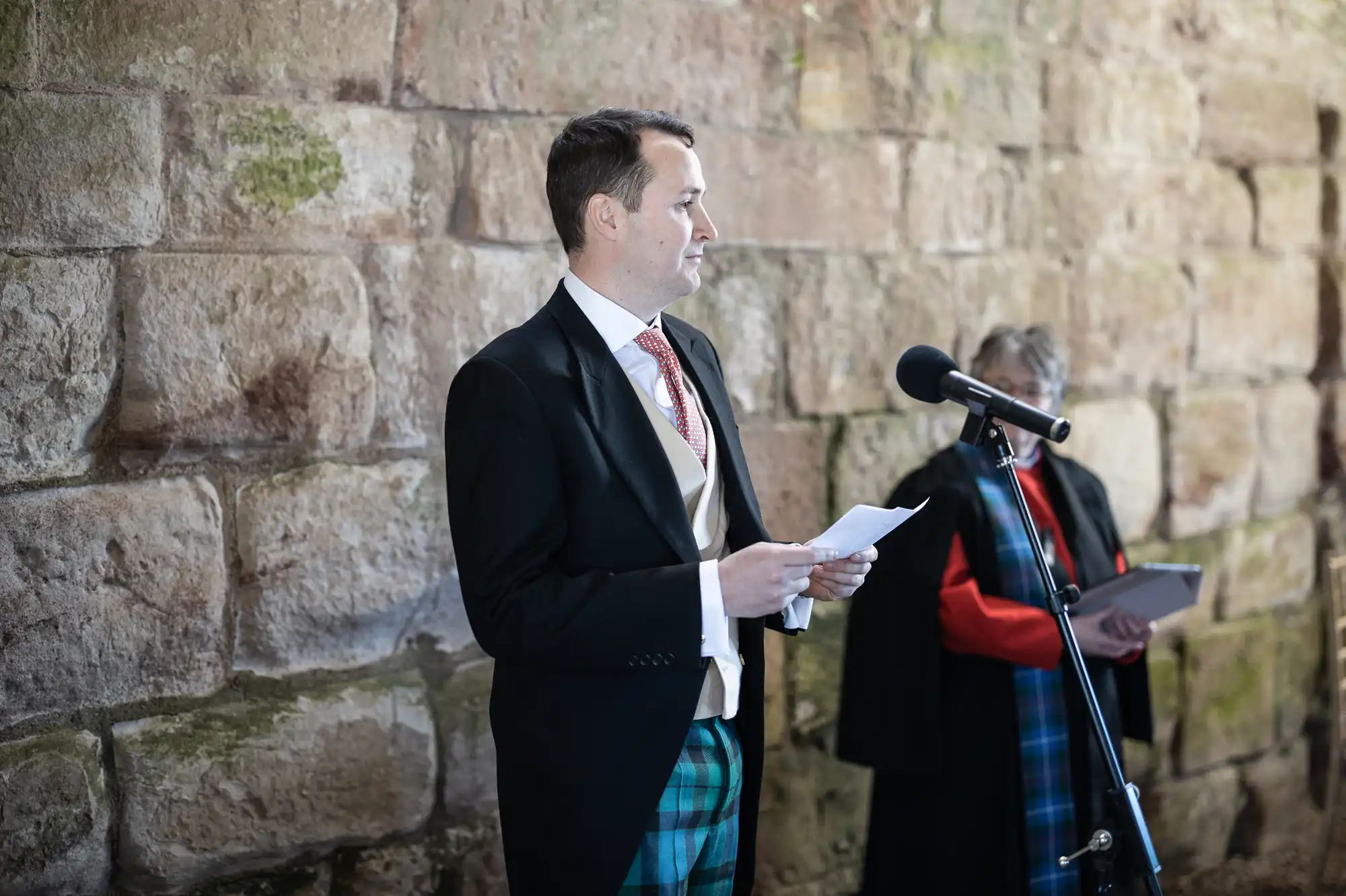 A man in formal attire speaks into a microphone while holding papers, standing in front of a stone wall. Another person is seen holding a book in the background.